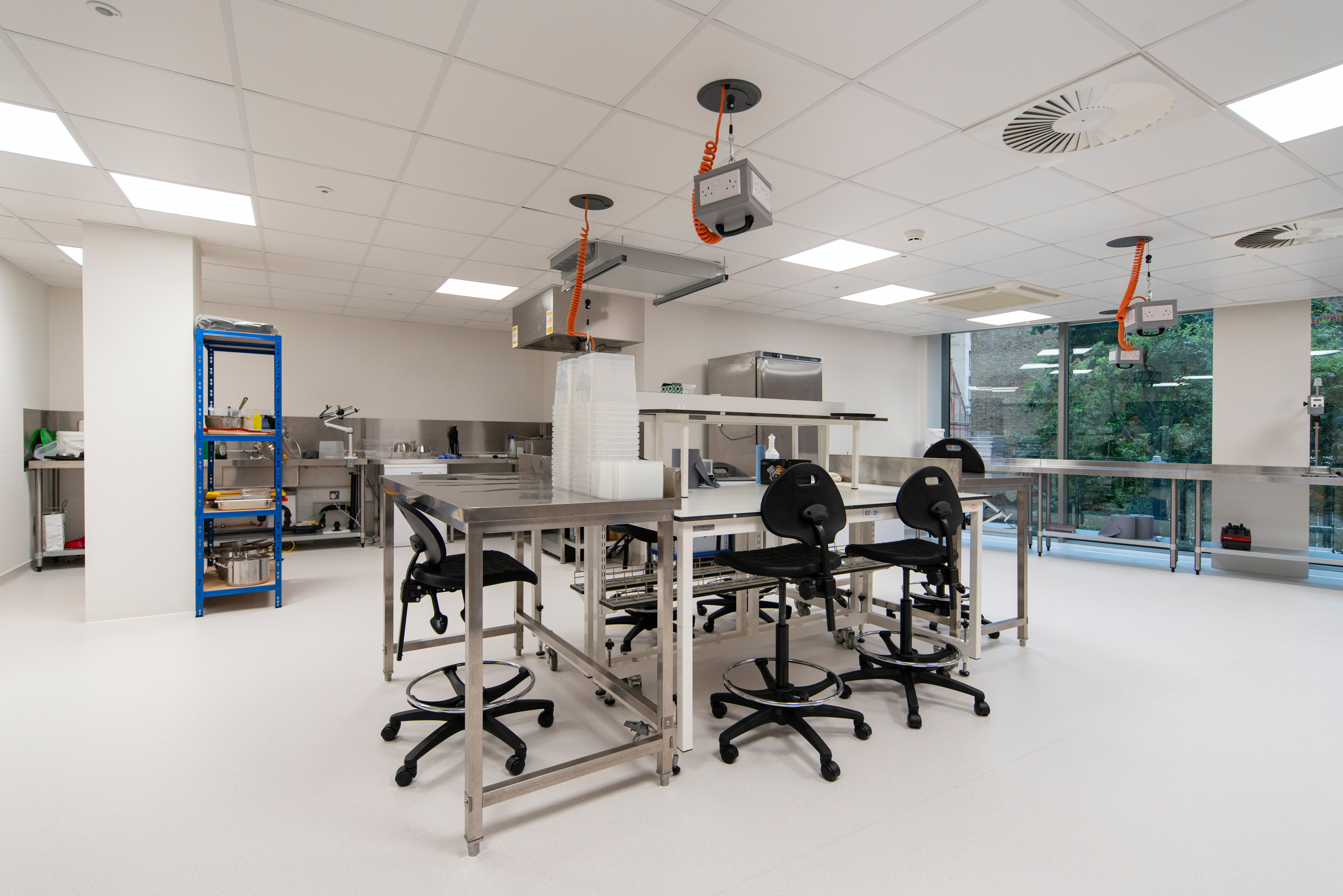 Modern commercial lab with stainless steel workstations, black swivel stools, and large windows allowing natural light.