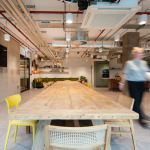 Modern office interior featuring a long wooden table surrounded by chairs, with a green sofa in the background, exposed ductwork, and a television mounted on a