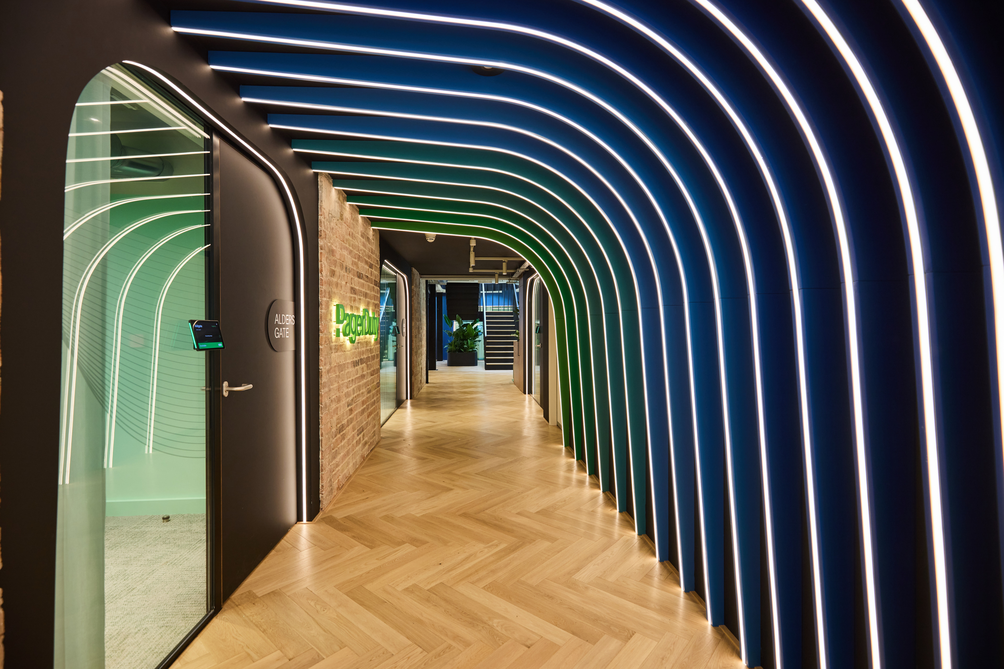 A modern hallway featuring curved, illuminated walls in shades of blue and green, with a wooden herringbone floor and a visible sign for "PagerDuty.
