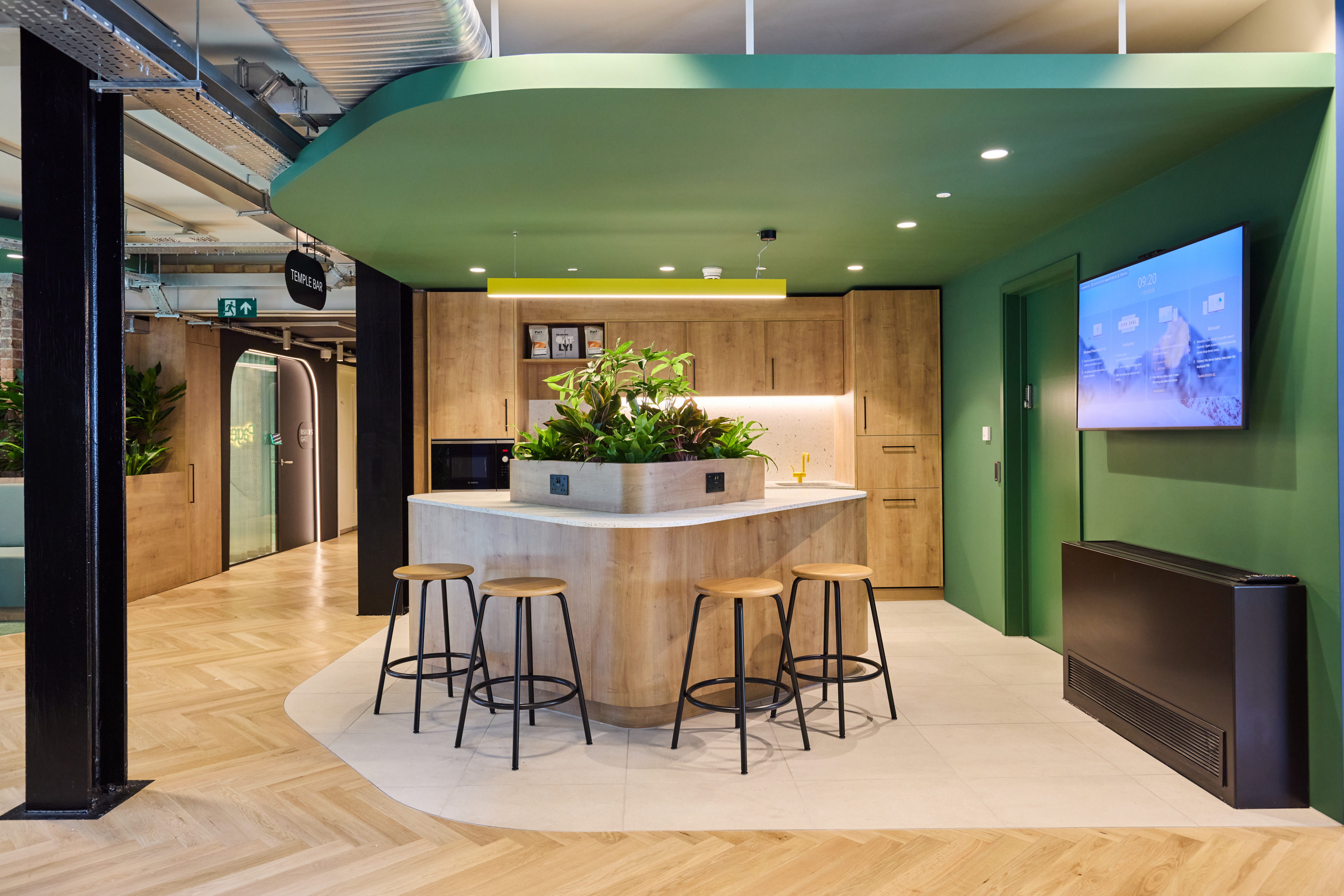 Modern kitchen area featuring a wooden island with a plant centerpiece, four stools, and a wall-mounted screen, surrounded by green and wooden accents.