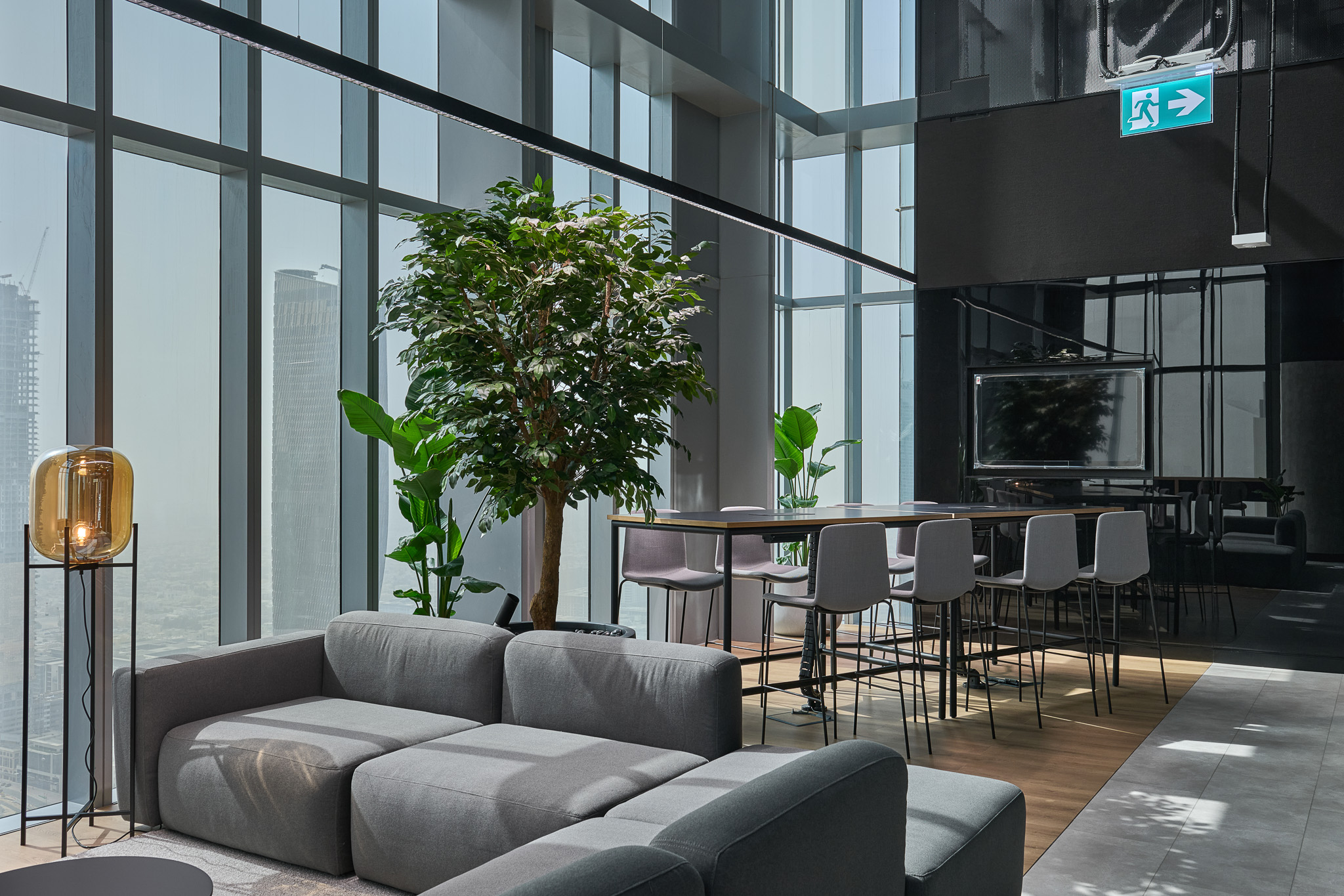 Modern office interior featuring a gray sectional sofa, a decorative plant, a standing lamp, and a dining table with chairs, all set against large windows with