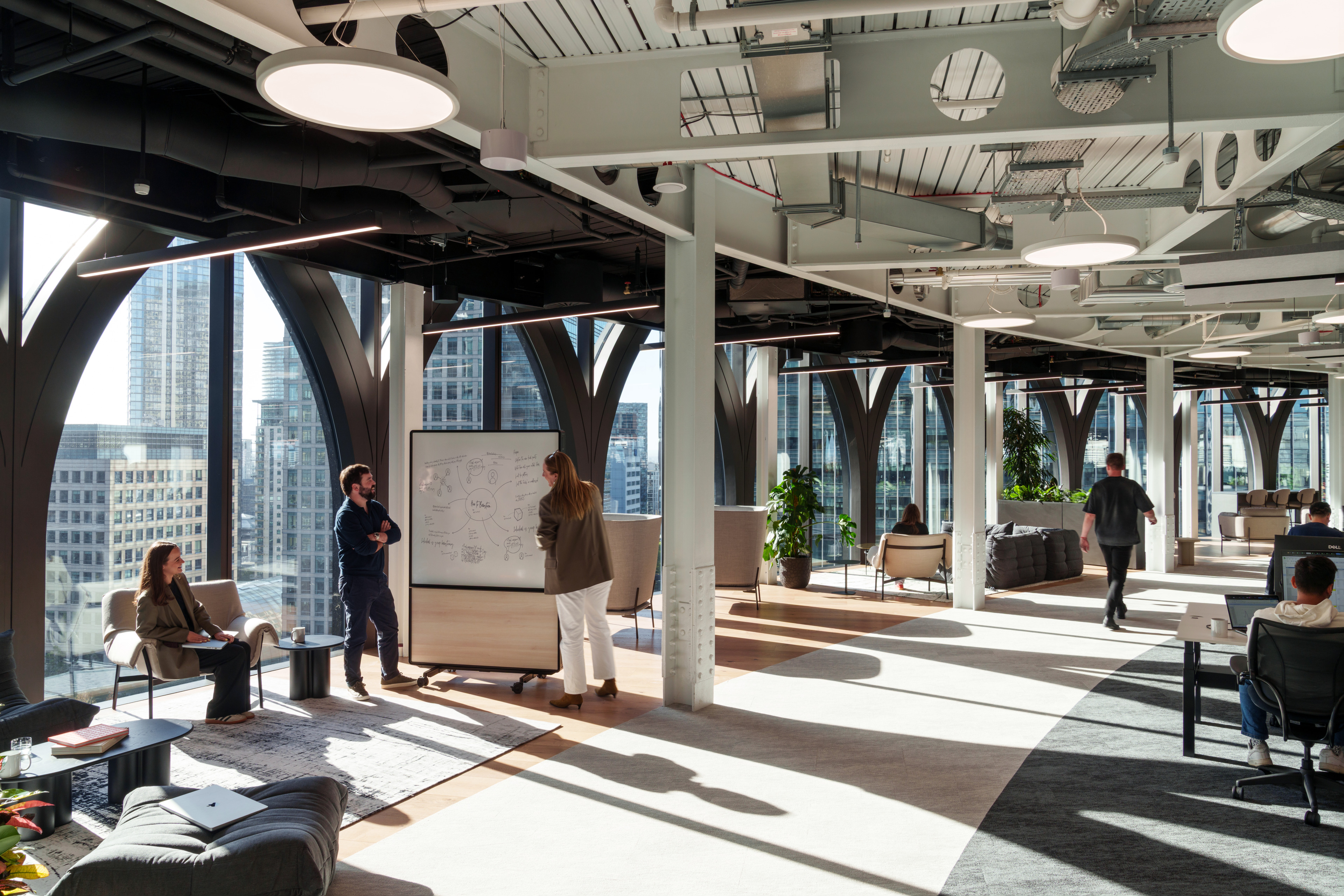 Modern office interior with large windows, featuring a group of people collaborating around a whiteboard, comfortable seating areas, and greenery.