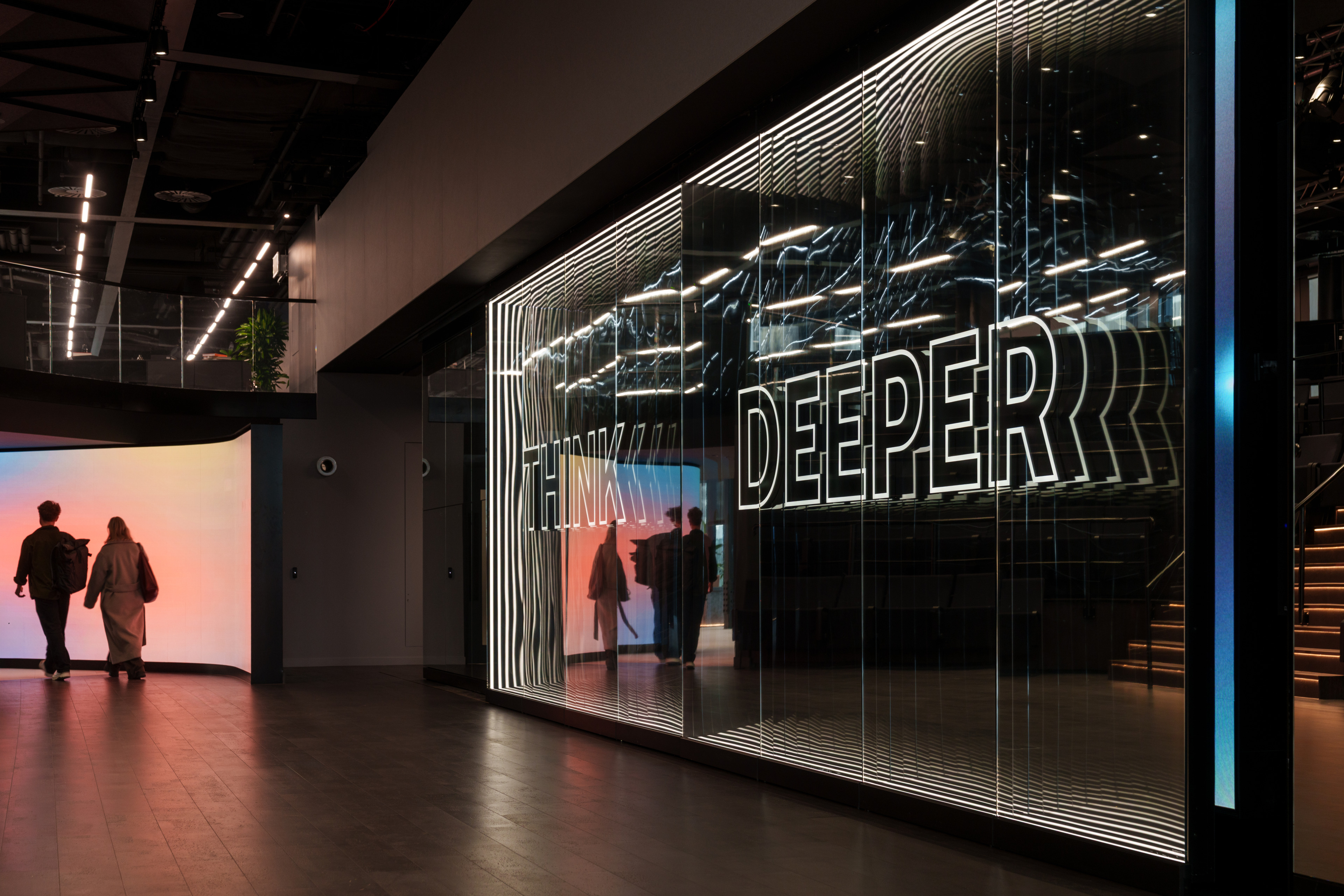 Interior view of a modern space featuring a large glass wall with the words "THINK DEEPER" illuminated, and two figures walking towards a colorful display in th
