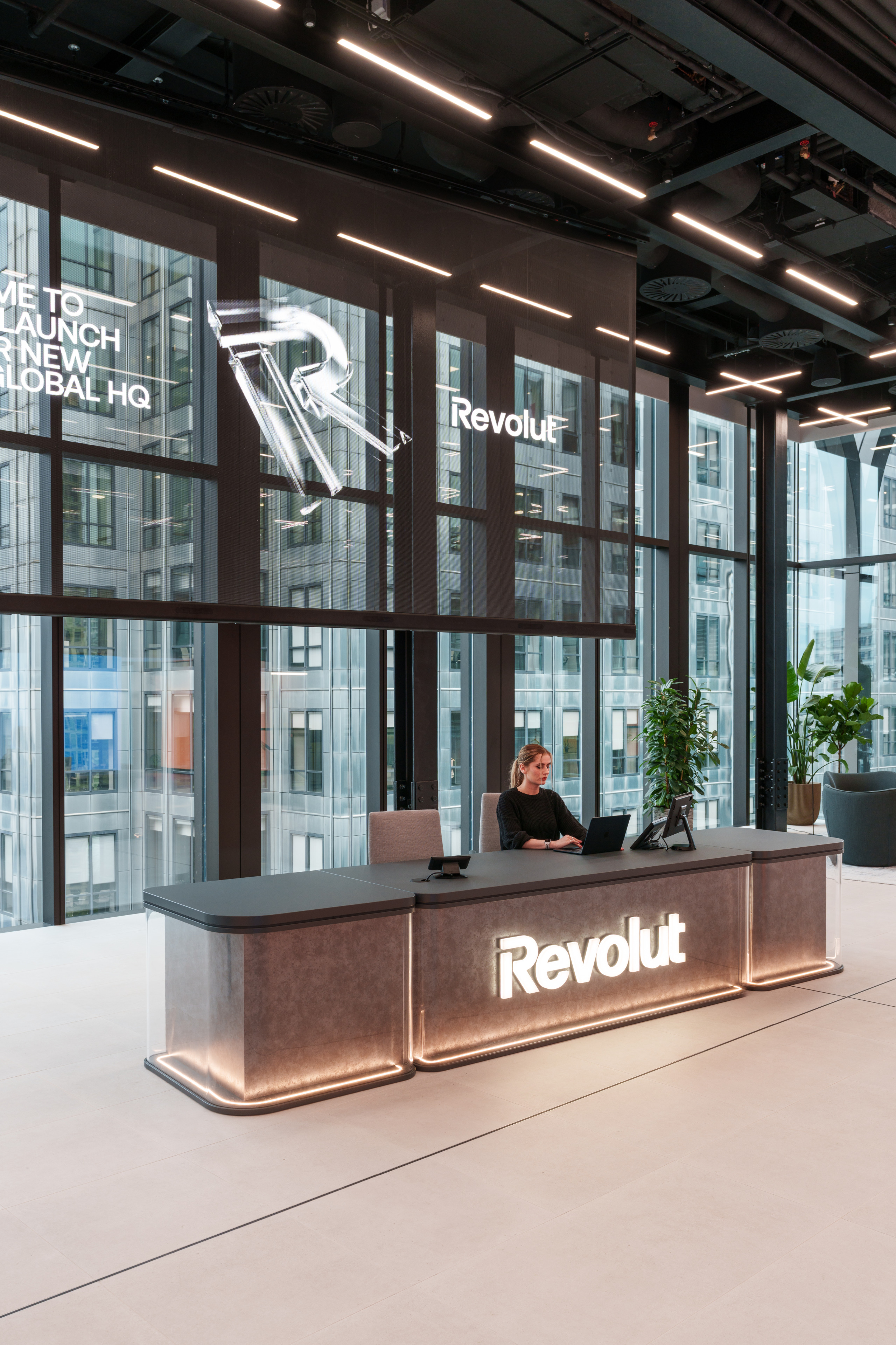 Reception area of Revolut's global headquarters, featuring a woman at a desk with a laptop, illuminated signage, and large windows showcasing the building's ext