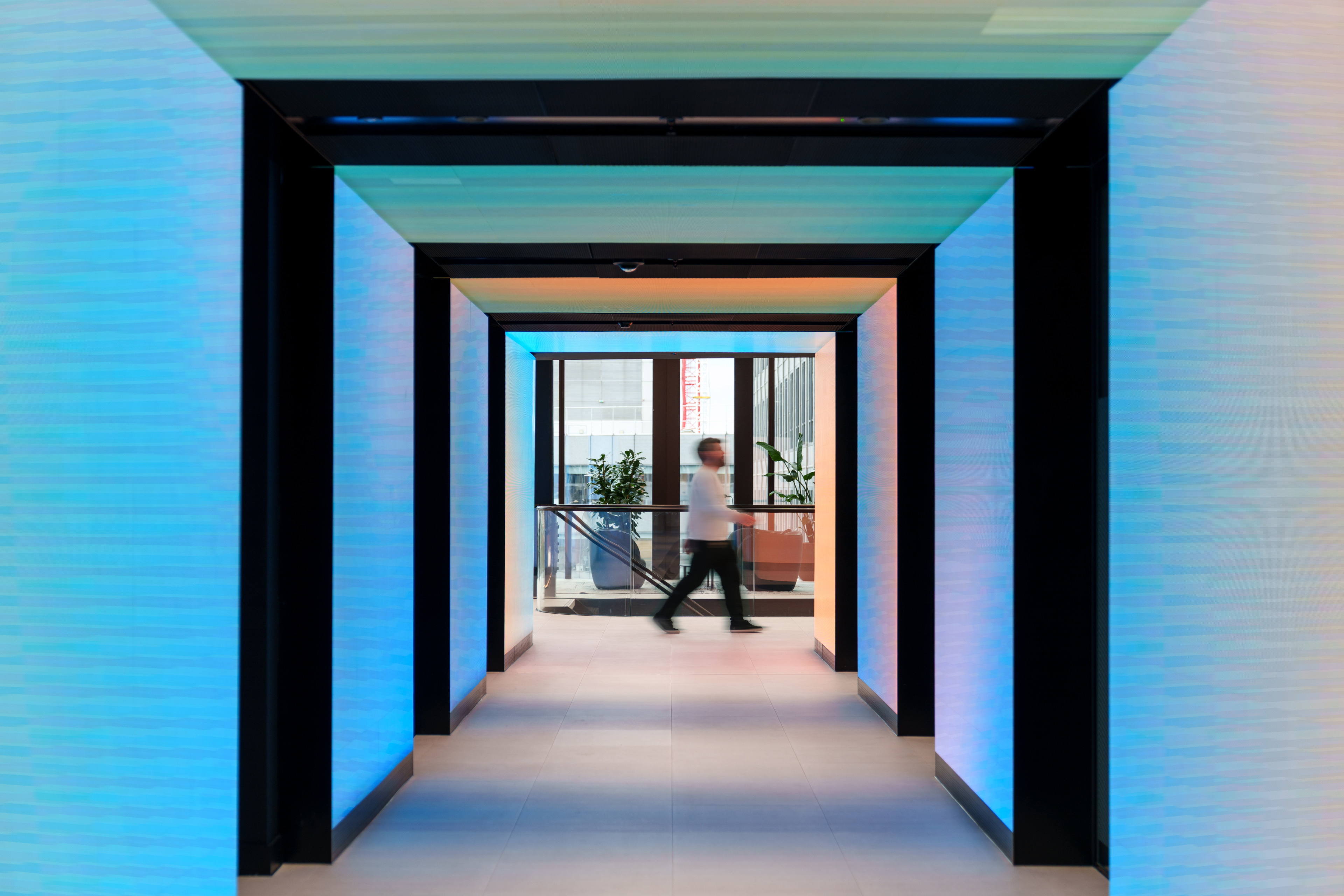 A person walks through a brightly lit corridor with colorful walls and a view of plants and glass railings at the end.