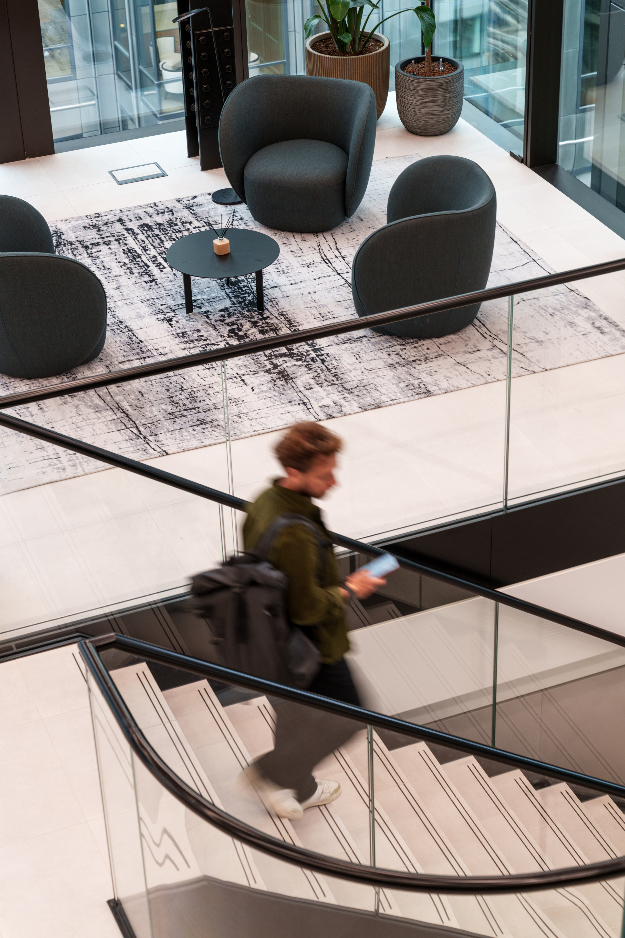 A person walks down a staircase with a black backpack, passing a seating area with two gray chairs and a small black table on a patterned rug.