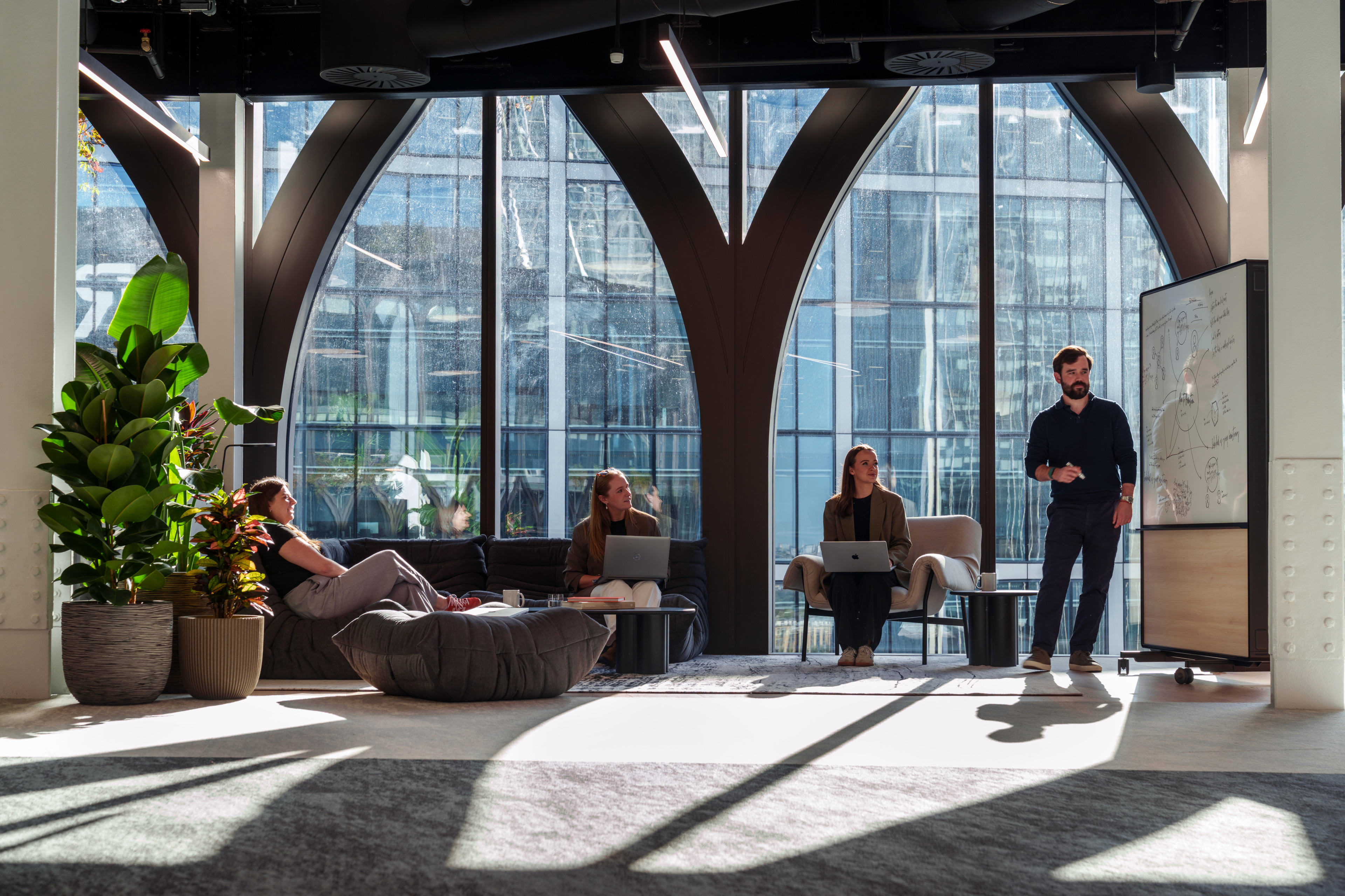 A man presents ideas on a whiteboard to three colleagues seated in a modern office space with large windows and plants.