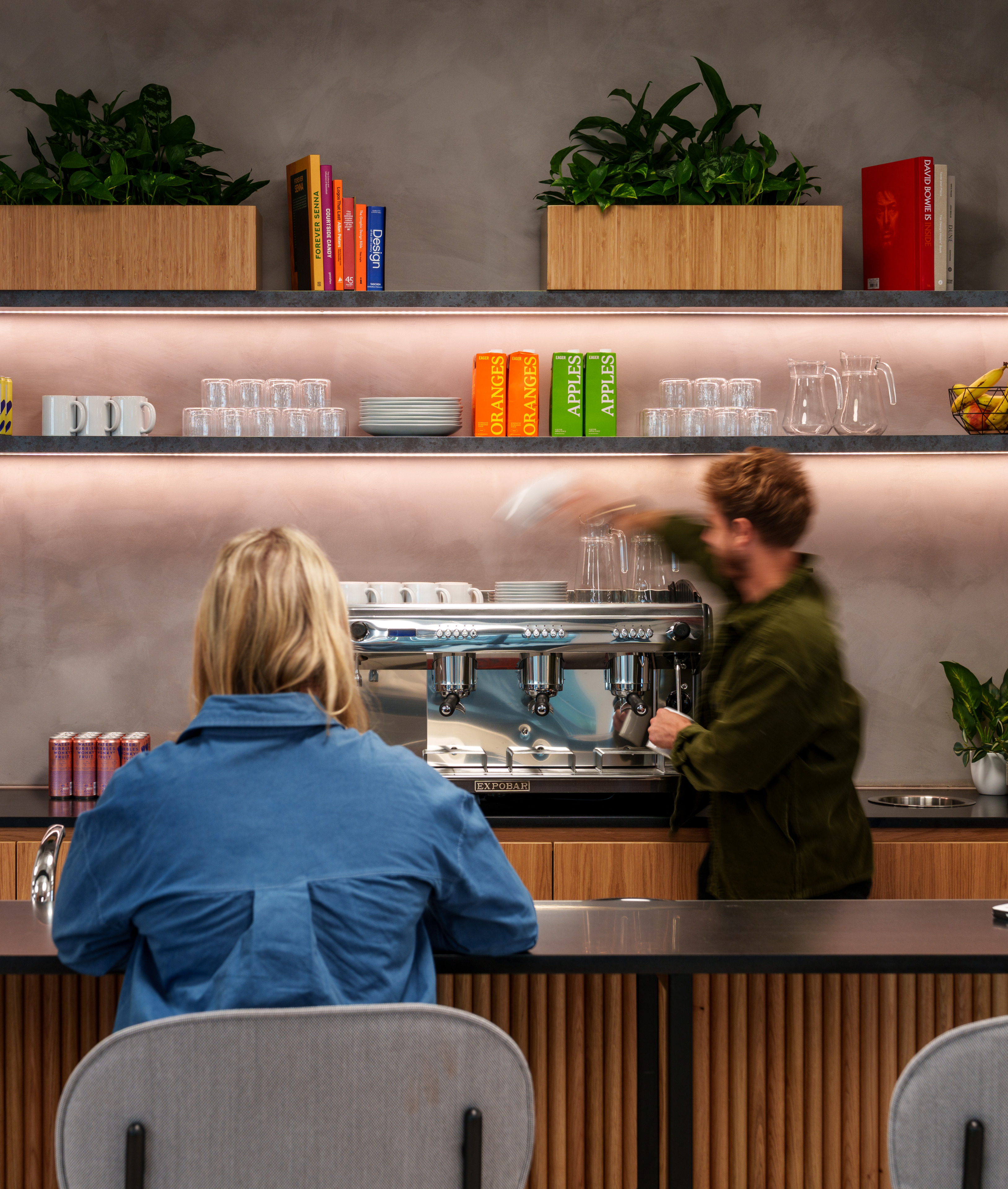 A person in a blue shirt sits at a bar counter while a barista prepares coffee behind an espresso machine, with shelves displaying plants, mugs, and colorful bo