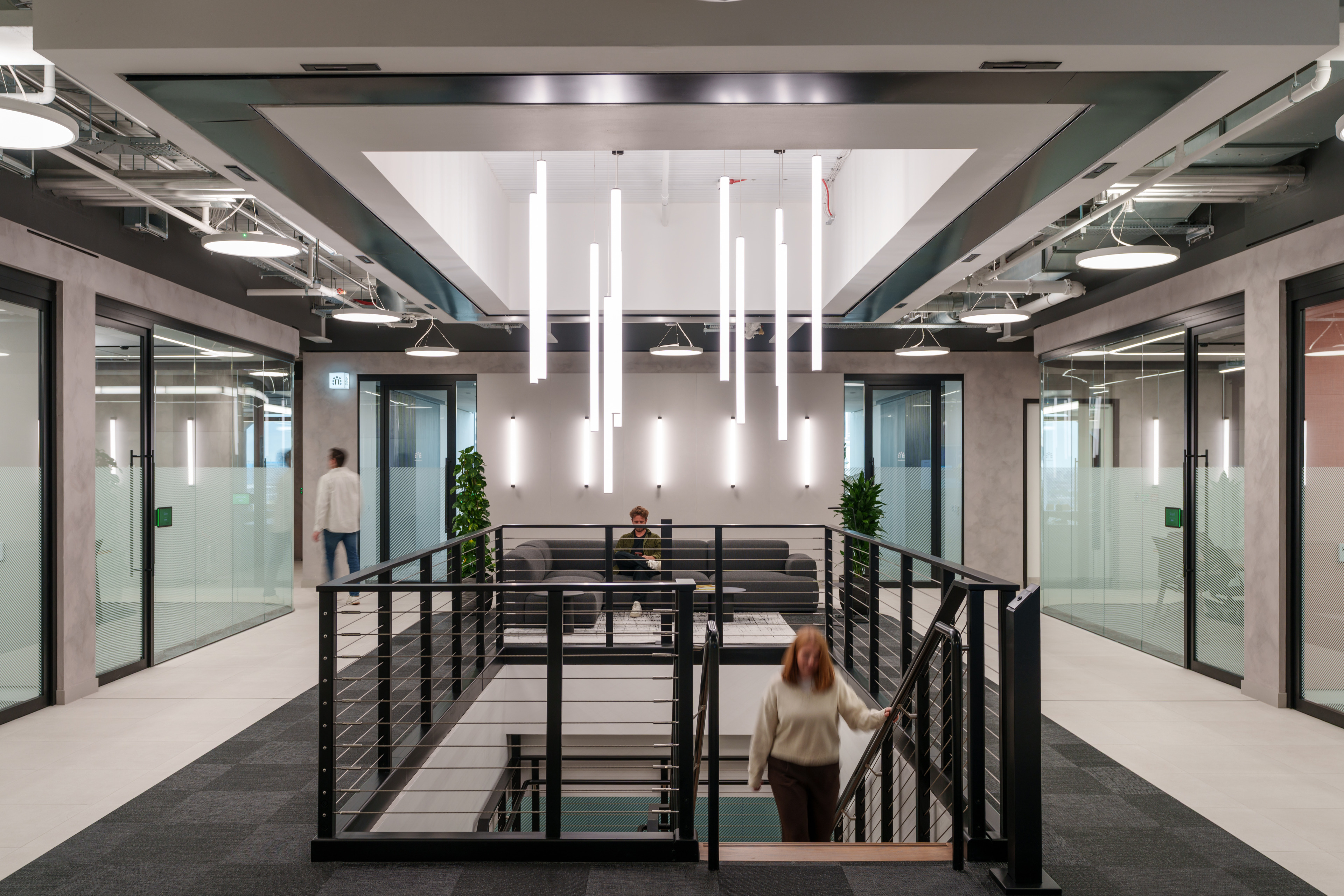 Modern office interior featuring a staircase, seating area, and glass-walled meeting rooms, illuminated by hanging light fixtures.