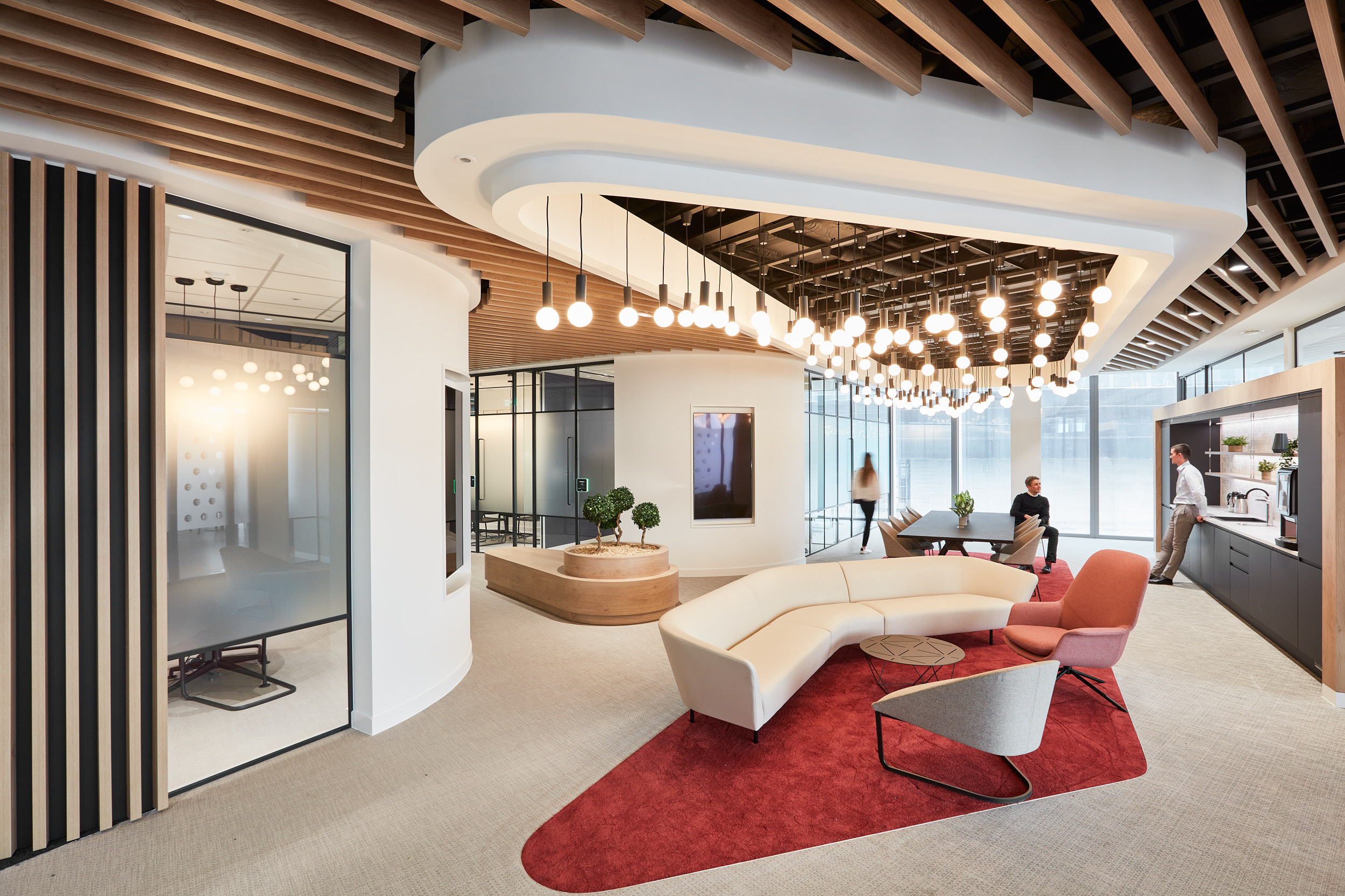 Modern office interior featuring a curved white sofa, a red area rug, and a wooden ceiling with hanging lights, with people engaged in conversation and a kitche