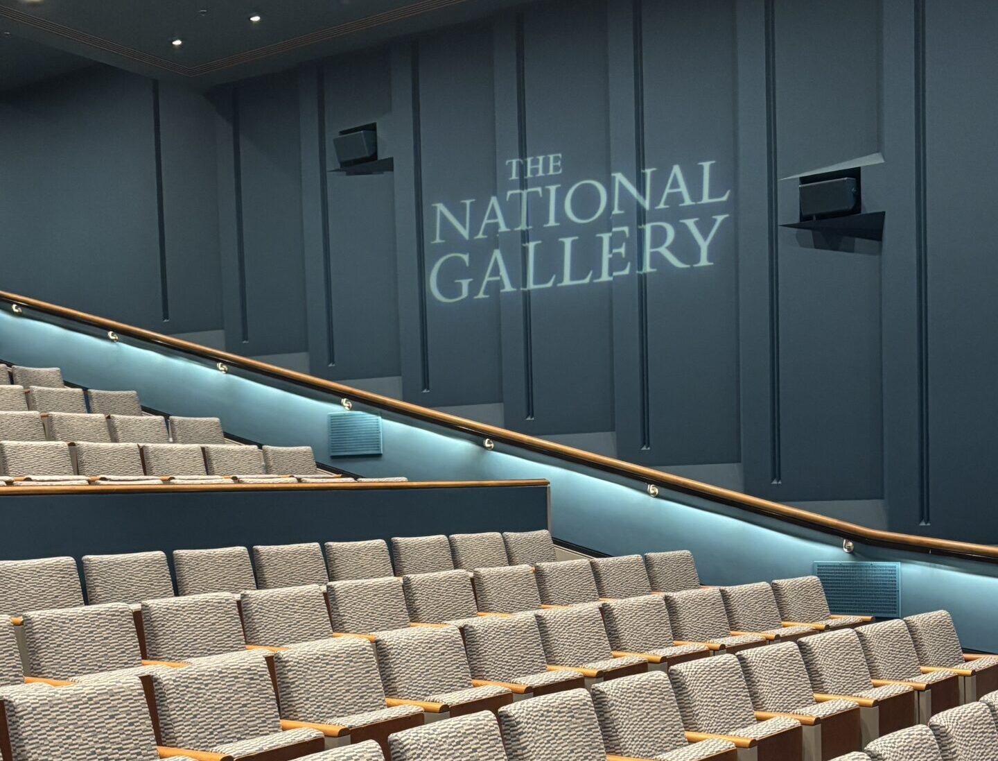 Seating area in a theater with a projection of "The National Gallery" on the wall.