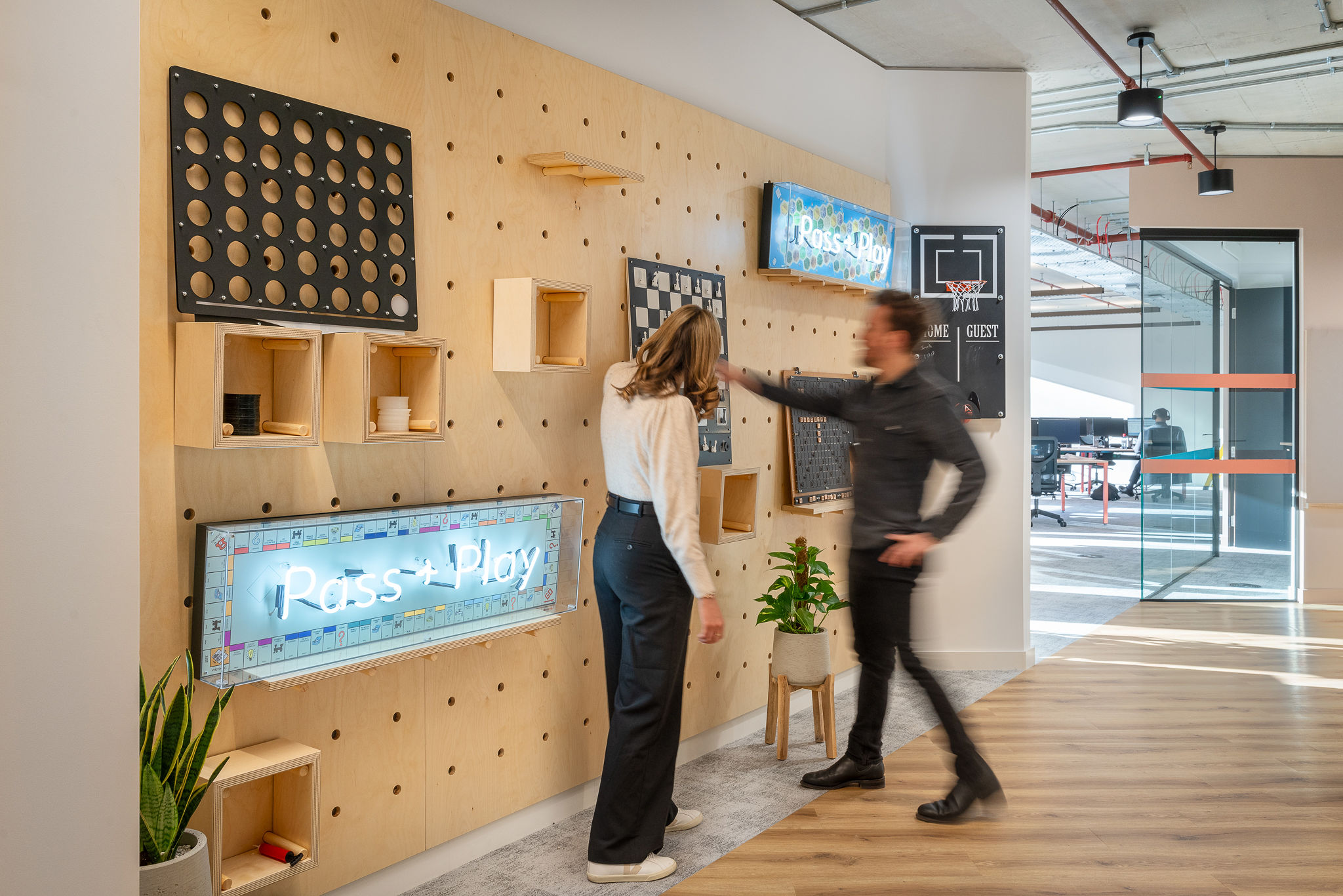 A woman and a man interact with a game wall featuring various board games and a neon sign that reads "Pass + Play" in a modern office space.
