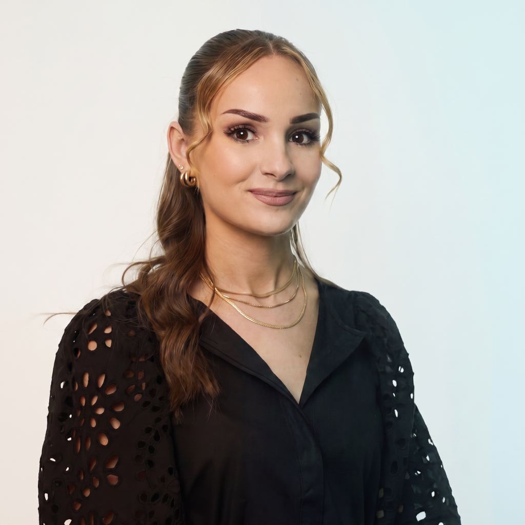 A woman with long, wavy hair wearing a black blouse with intricate cut-out designs and layered gold necklaces, smiling against a light background.
