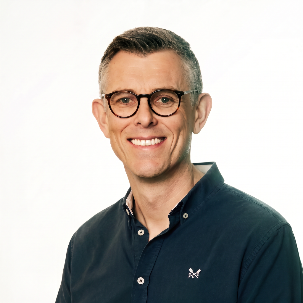 Man with short gray hair and glasses smiling, wearing a dark blue button-up shirt with a logo on the chest, against a light background.