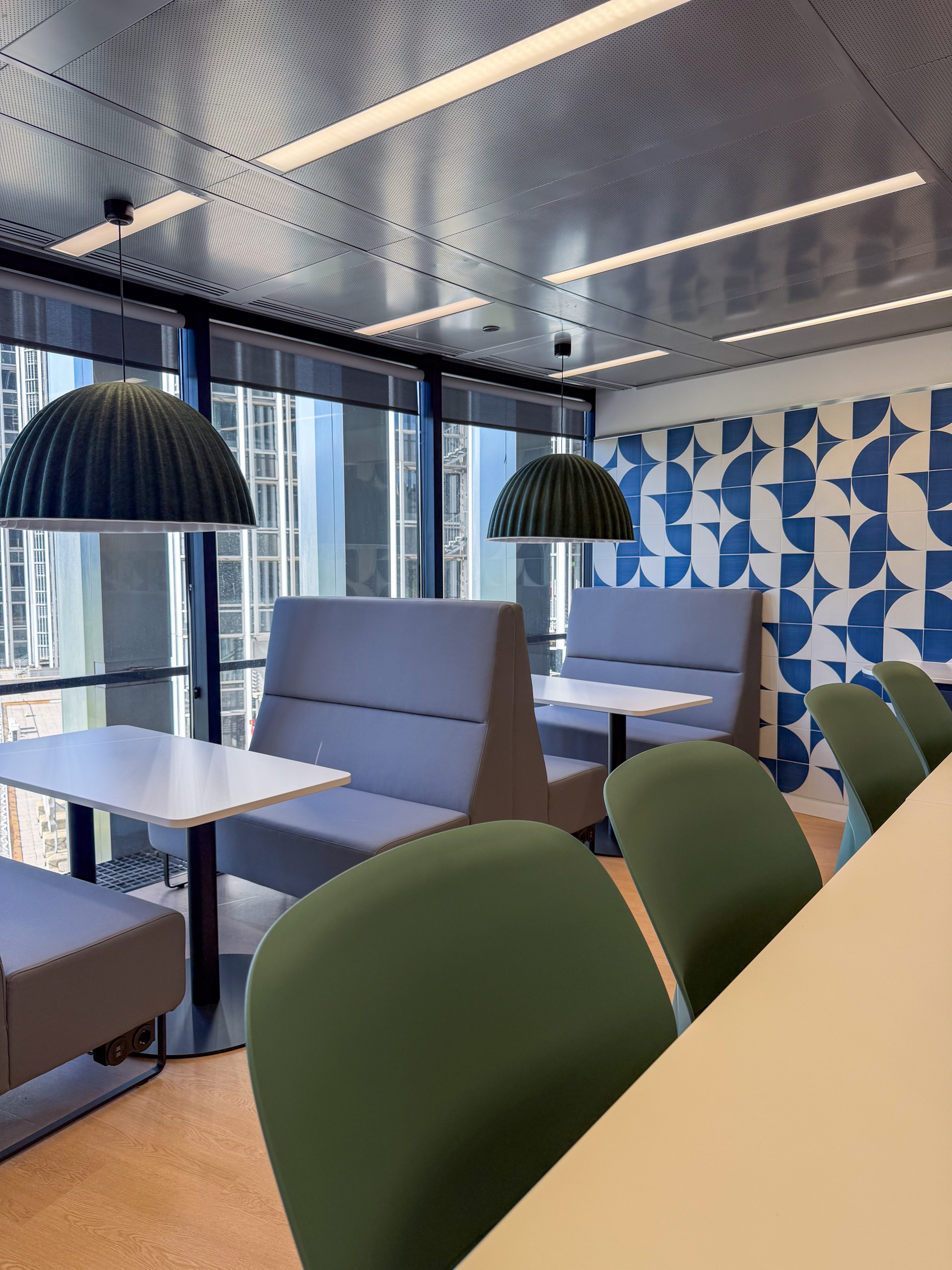 Modern office interior featuring gray seating booths, green chairs, and pendant lighting, with a patterned blue and white wall and large windows.