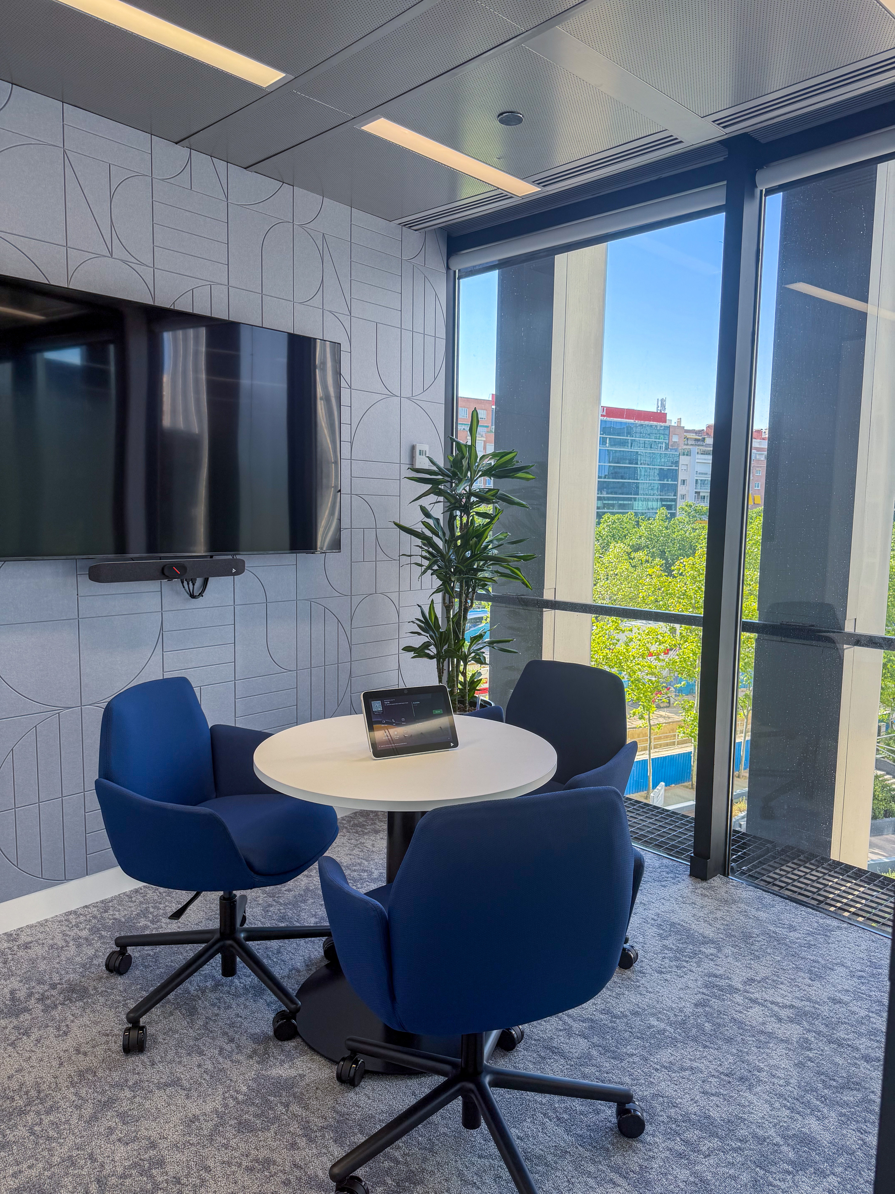 Modern meeting room featuring a round table with four blue chairs, a tablet on the table, a large screen on the wall, and large windows providing a view of the