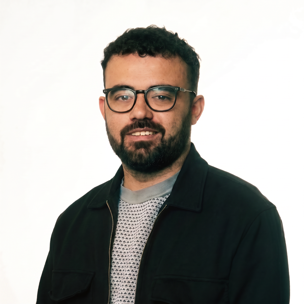 Man with curly hair and glasses smiling, wearing a dark jacket over a patterned sweater, against a white background.