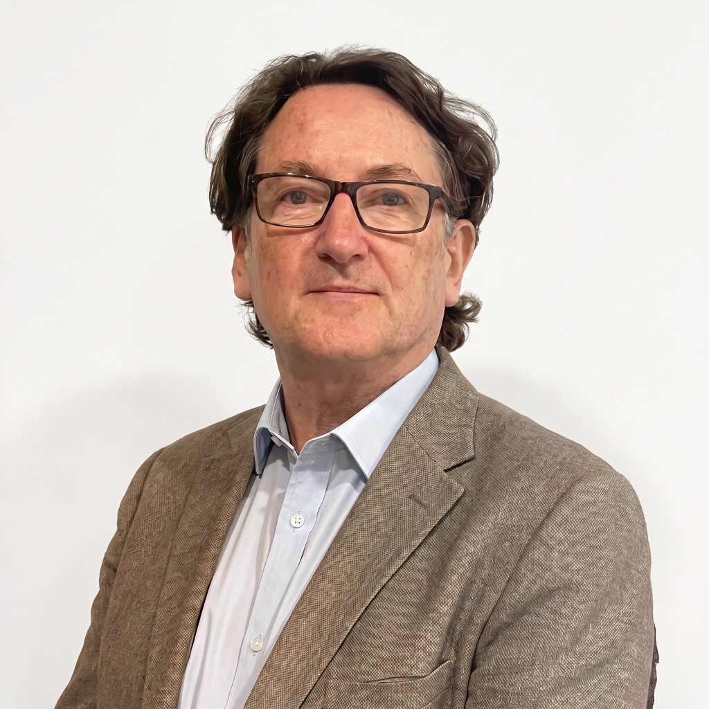 A middle-aged man with glasses and wavy hair, wearing a light blue shirt and a brown blazer, stands against a plain white background.