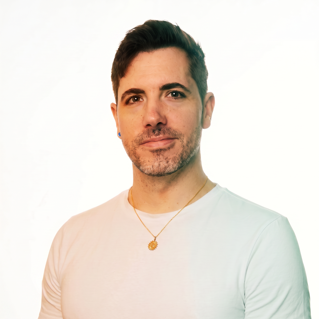 Man with short dark hair and a beard, wearing a white shirt and a gold necklace, against a light background.