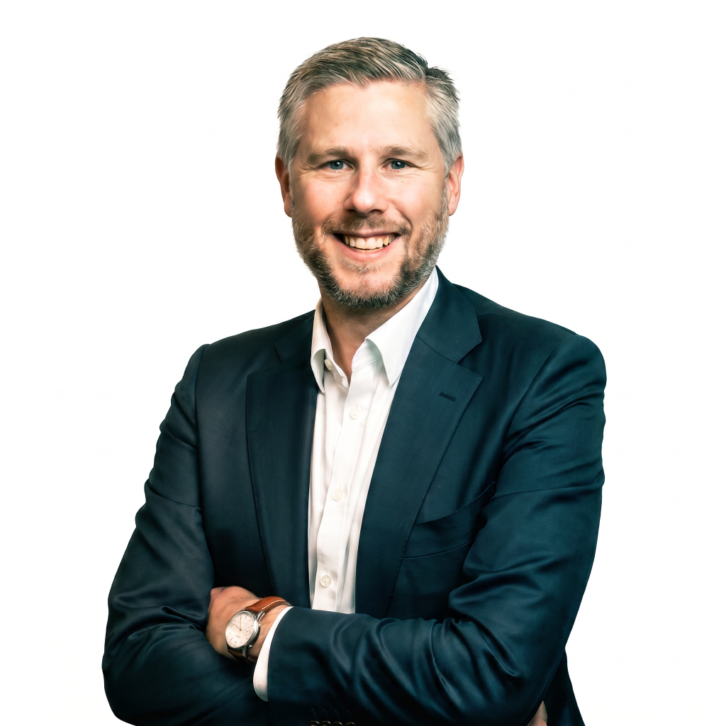 Smiling man with short gray hair and a beard, wearing a dark suit and white shirt, standing with arms crossed against a white background.