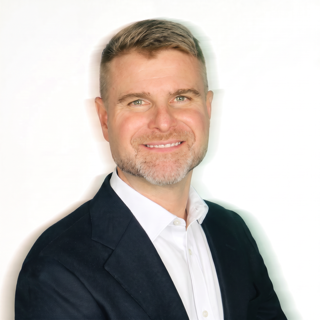 Portrait of a smiling man with short blonde hair and a beard, wearing a dark blazer over a white shirt, against a light background.