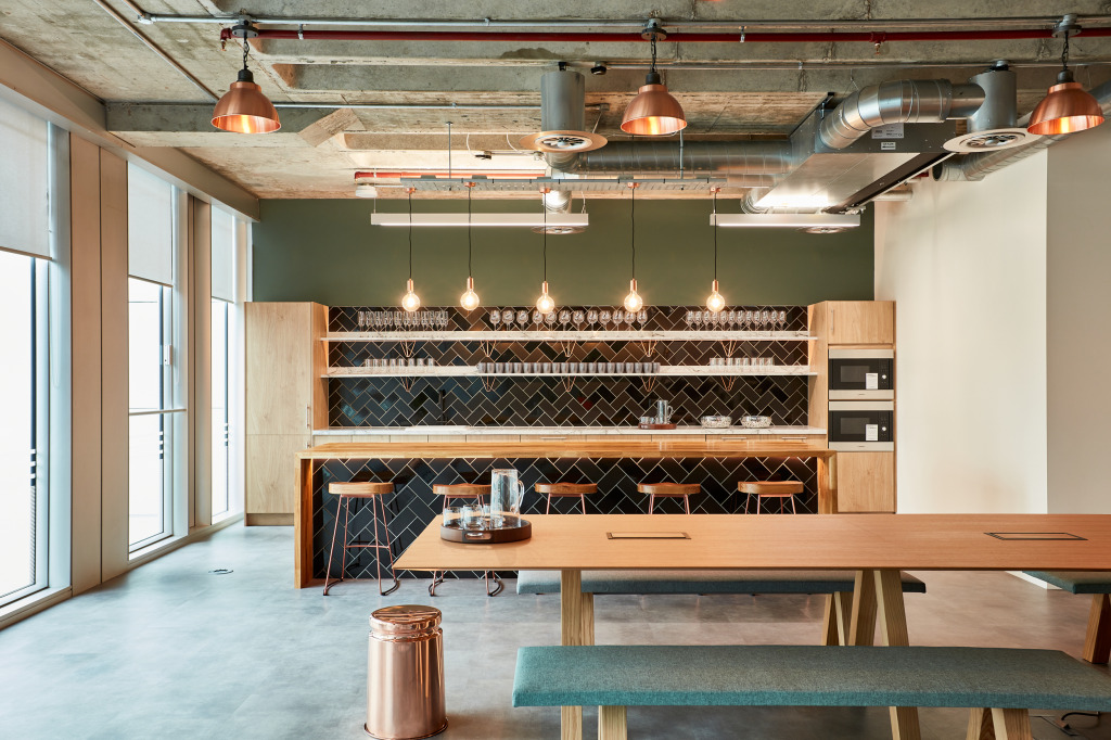 Modern kitchen area featuring a wooden bar with high stools, a long dining table, and shelves displaying glassware, set against a green accent wall and industri