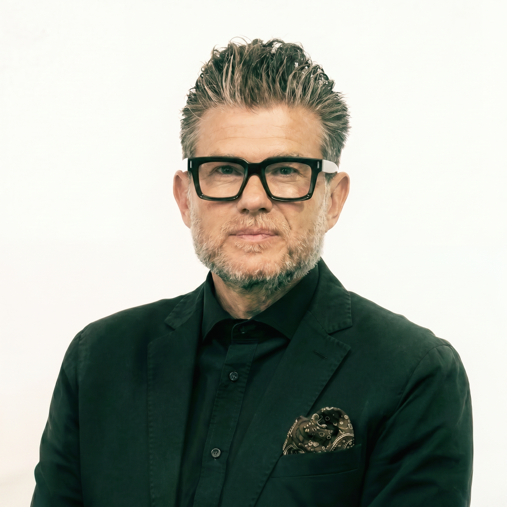 Man with styled hair and glasses, wearing a black blazer and shirt, with a patterned pocket square, against a white background.