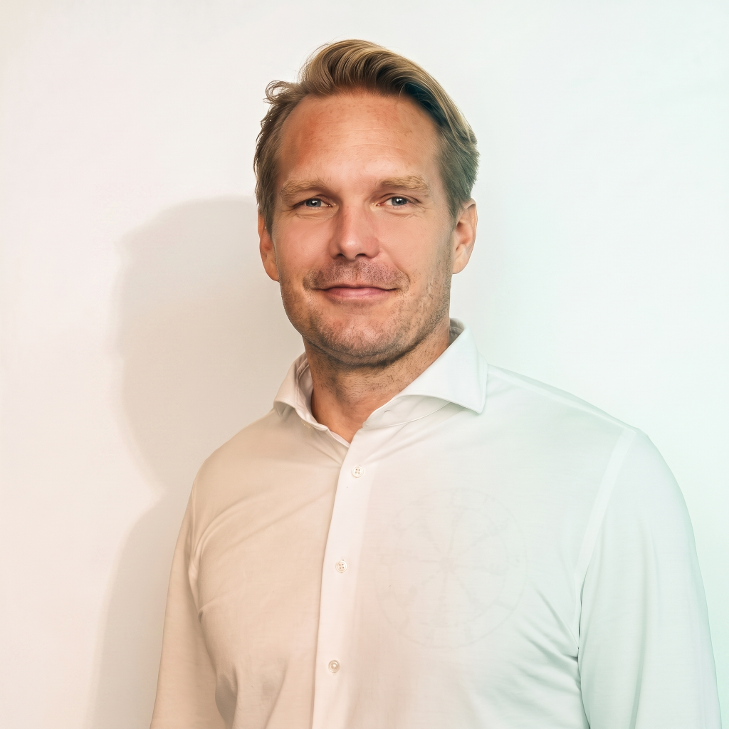 A man with short blond hair and a slight smile is wearing a white button-up shirt, standing against a light background.