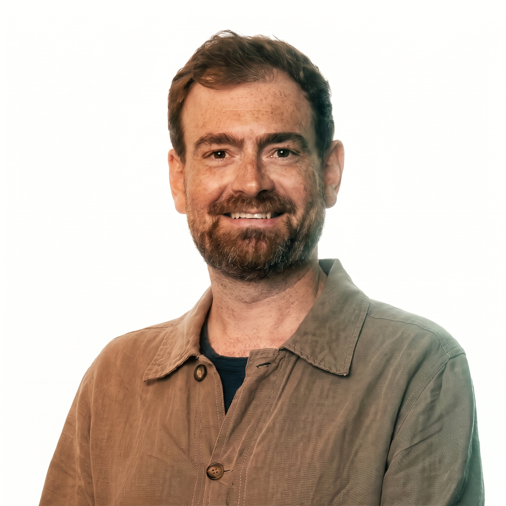 Man with a beard smiling, wearing a beige jacket over a dark shirt, against a white background.