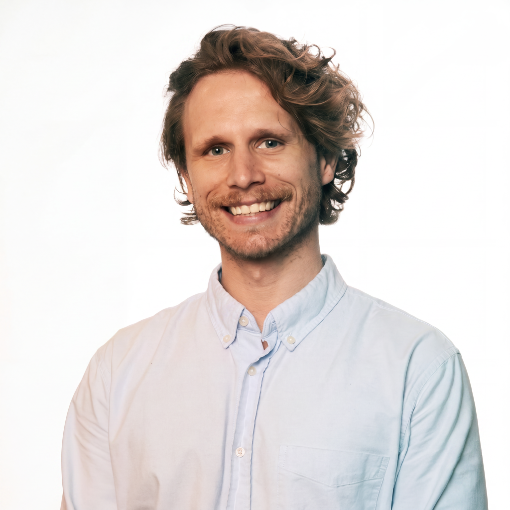 Smiling man with curly hair wearing a light blue button-up shirt against a white background.