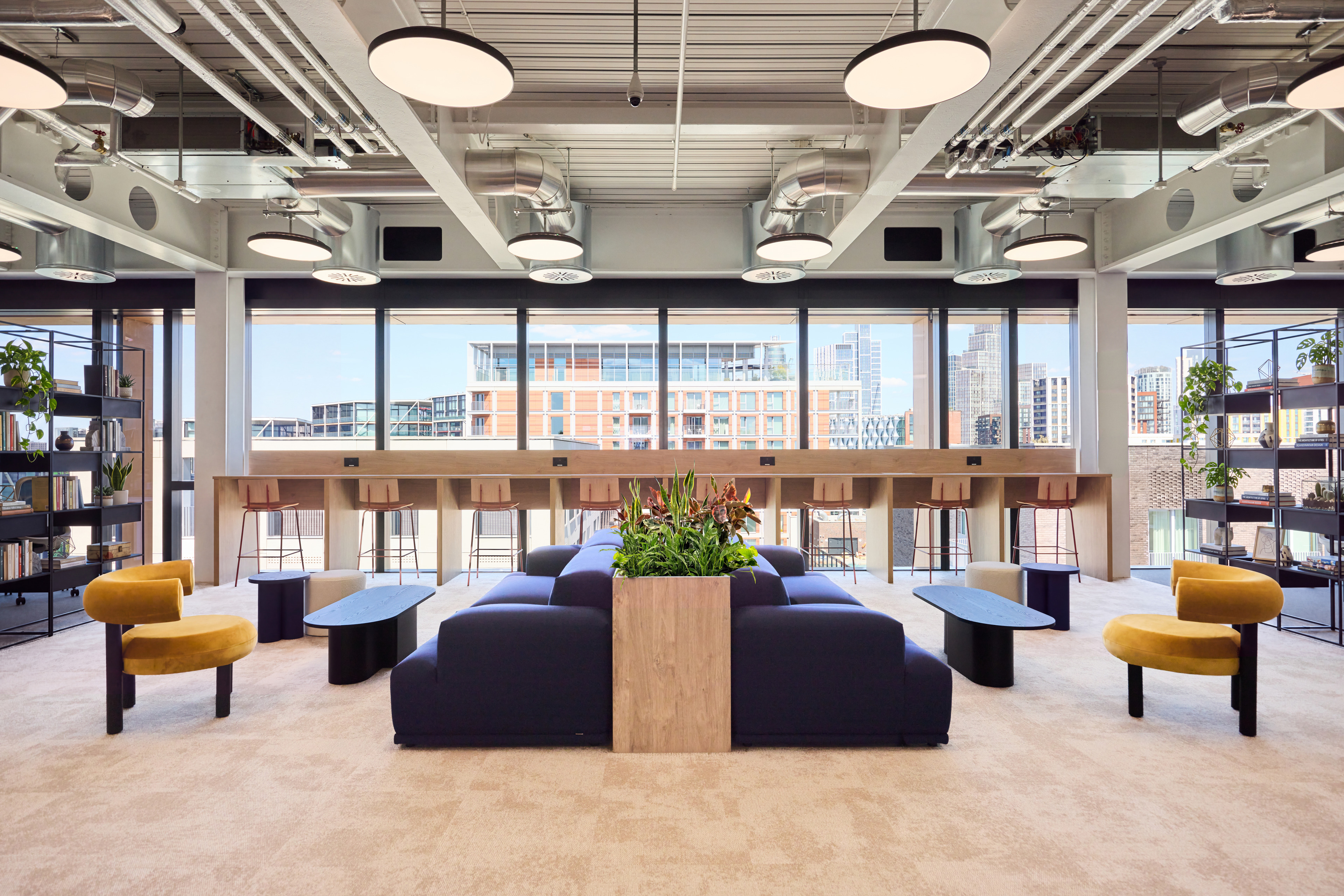 Modern office interior featuring a seating area with a large planter, bar-height tables, and shelves filled with plants and books, illuminated by circular ceili