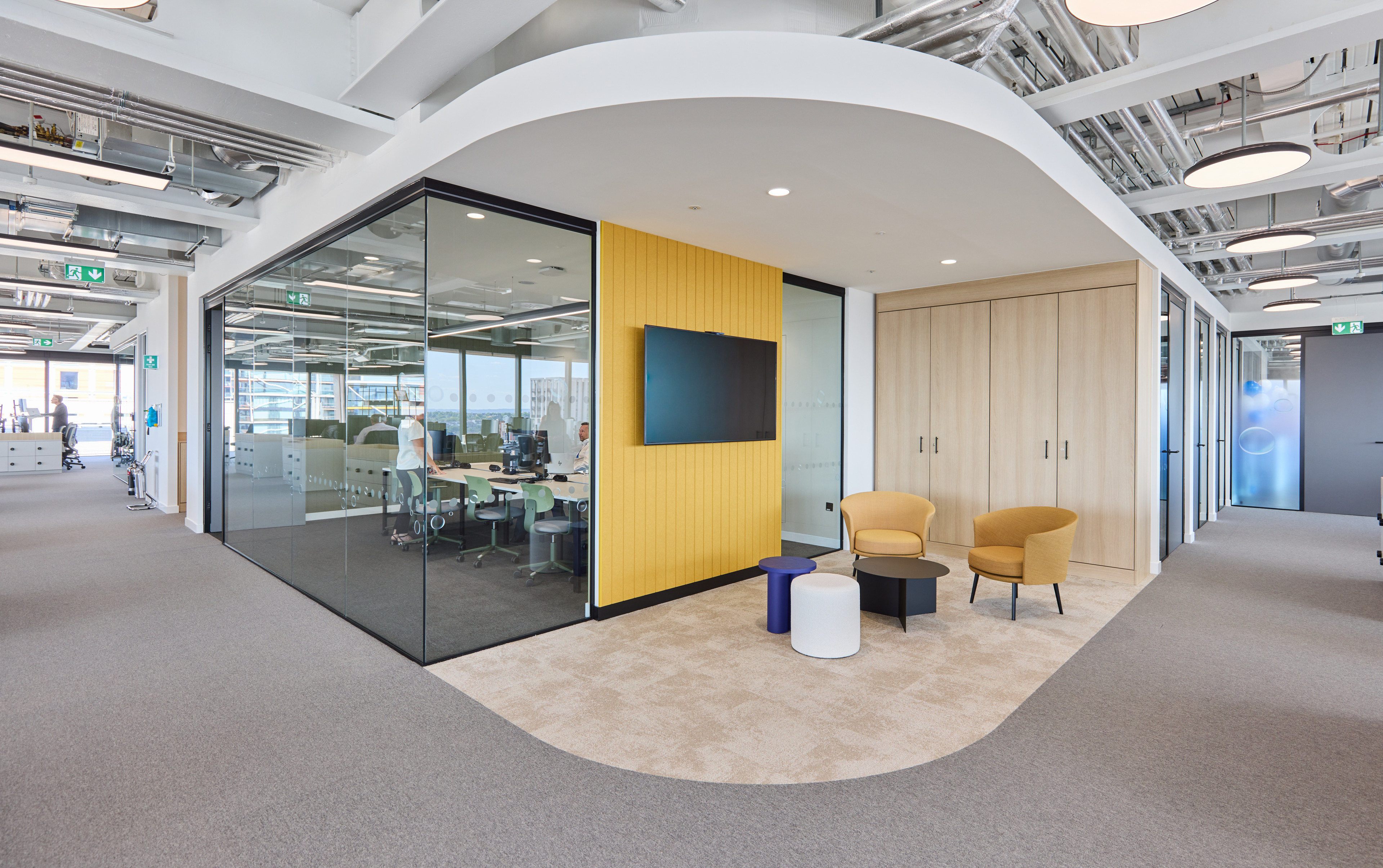 Modern office interior featuring a glass meeting room with a yellow accent wall, two yellow chairs, and a small table, alongside a workspace area with desks and