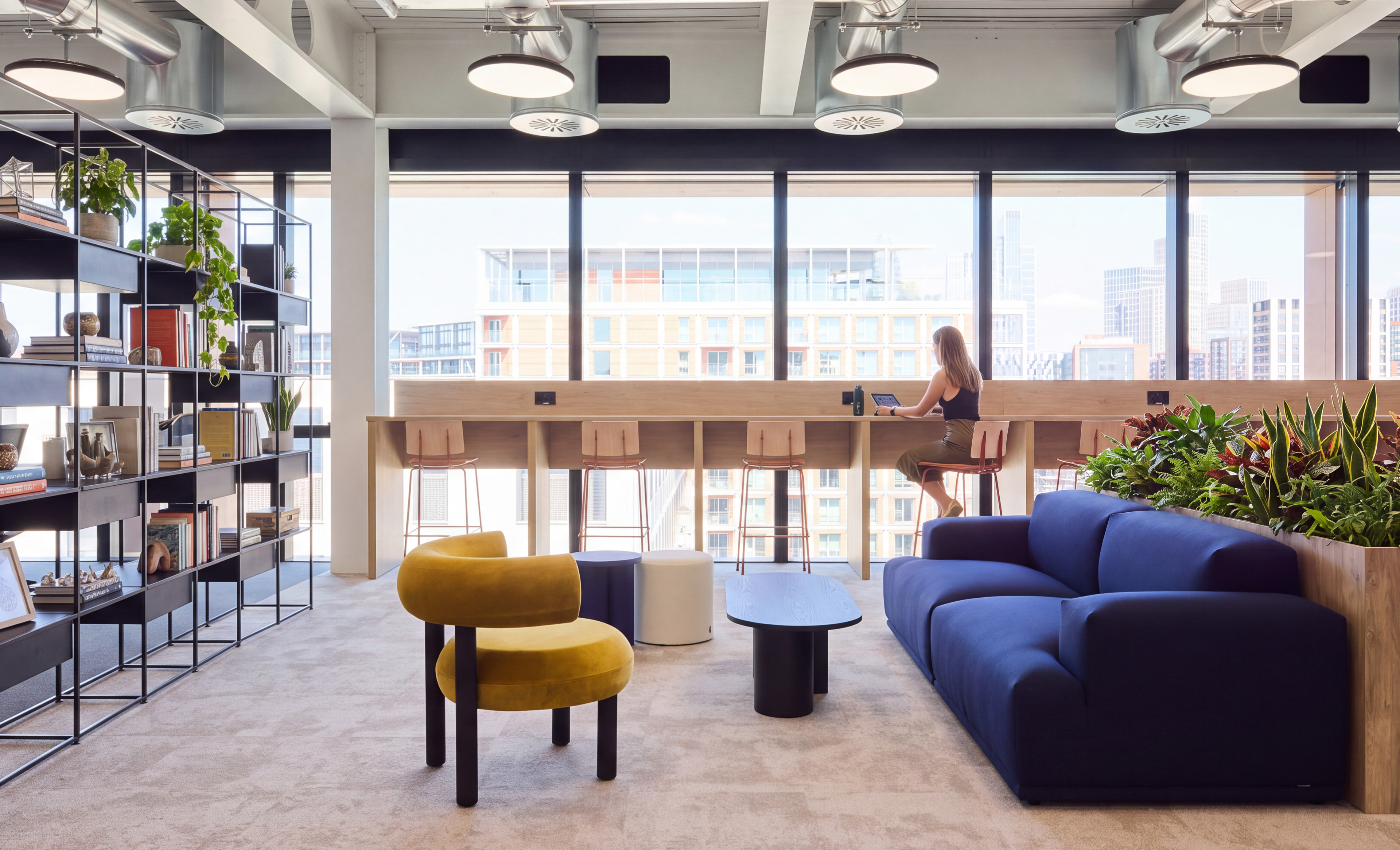 Modern office interior featuring a bookshelf, a blue sofa, and a woman working at a high table with large windows overlooking a cityscape.