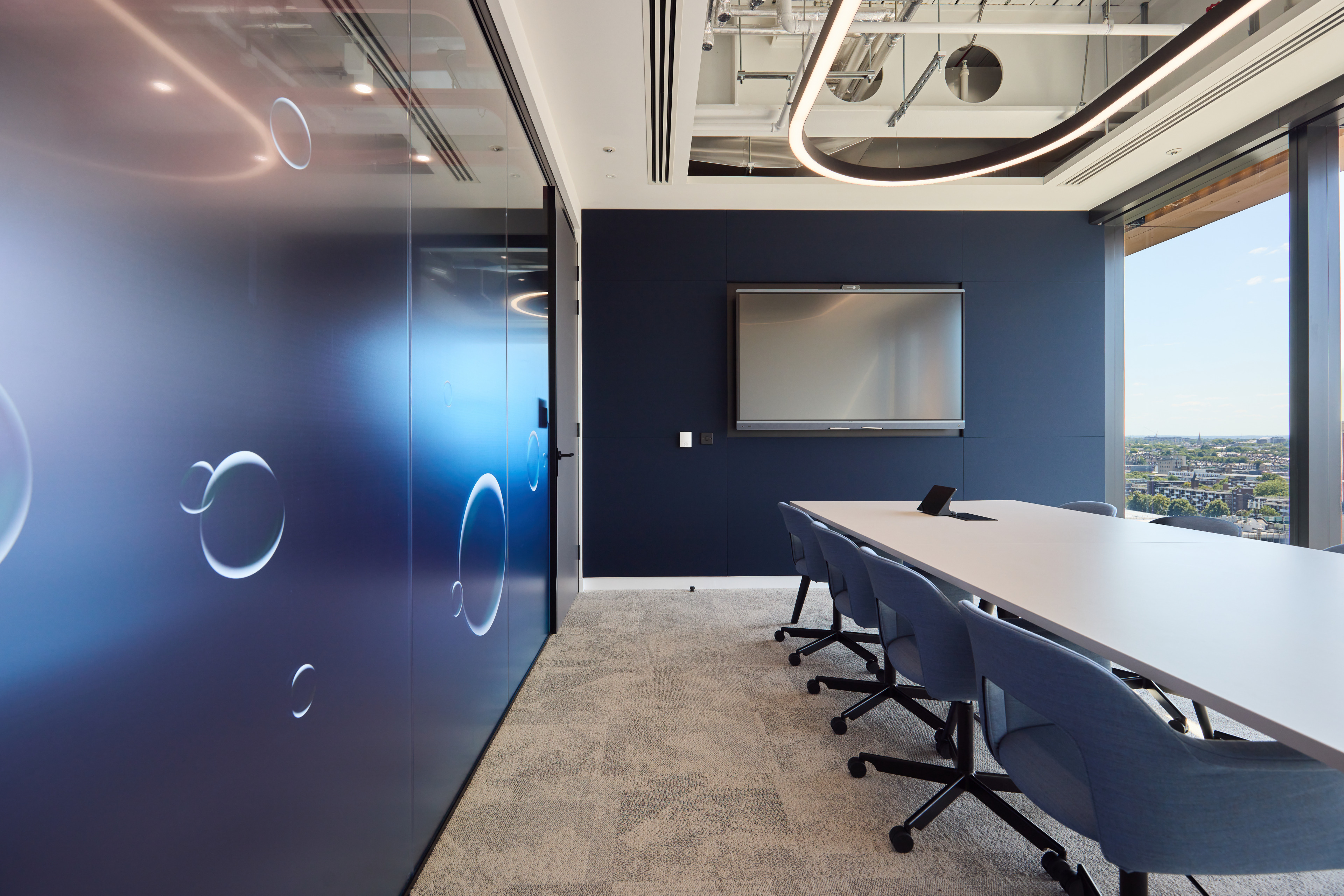 Modern conference room featuring a long white table, blue chairs, a wall with bubble designs, and a large screen mounted on the wall, with a view of the city vi