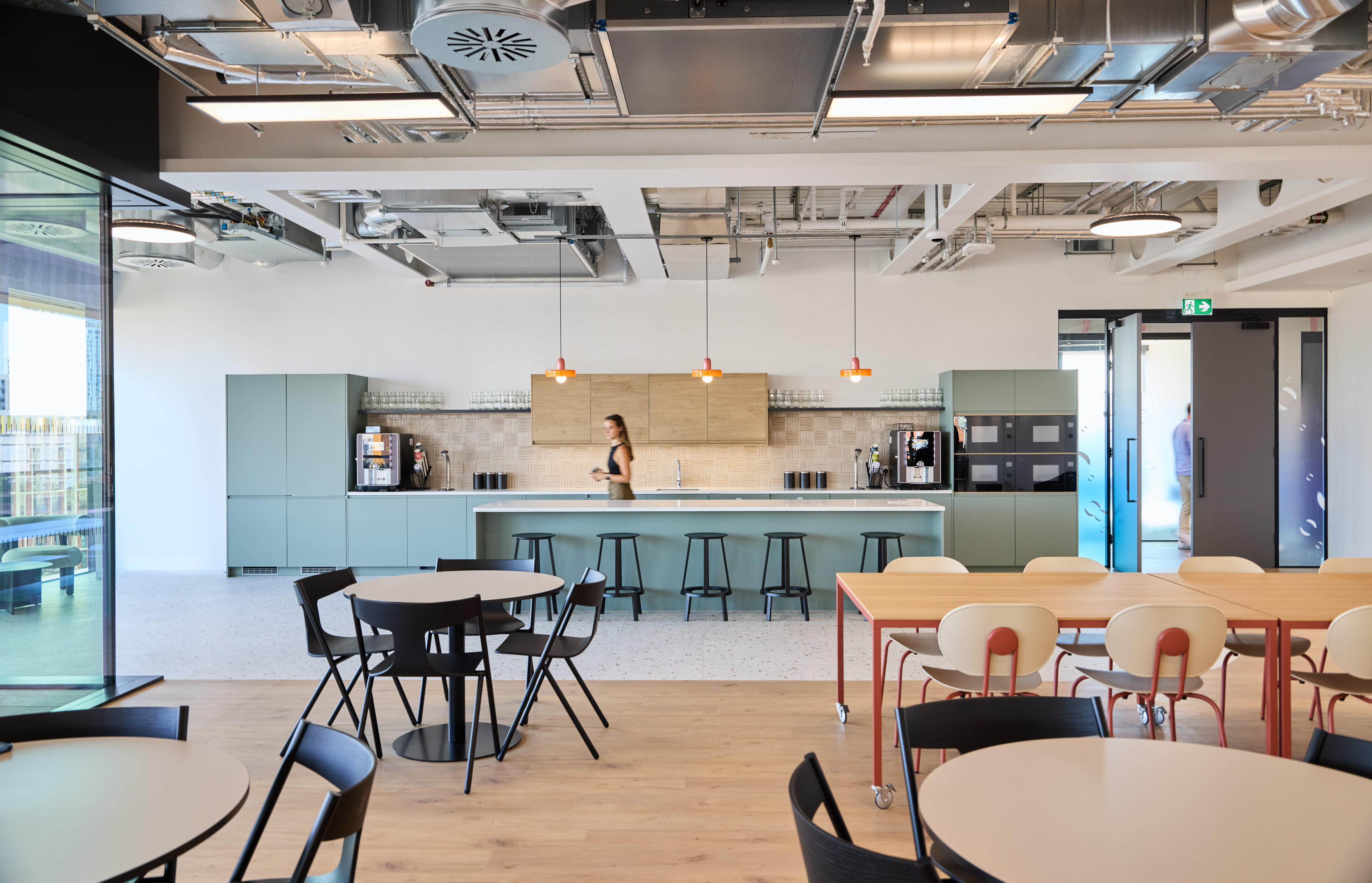 Modern office kitchen with a woman walking past a counter, featuring green cabinetry, coffee machines, and seating areas with round tables.