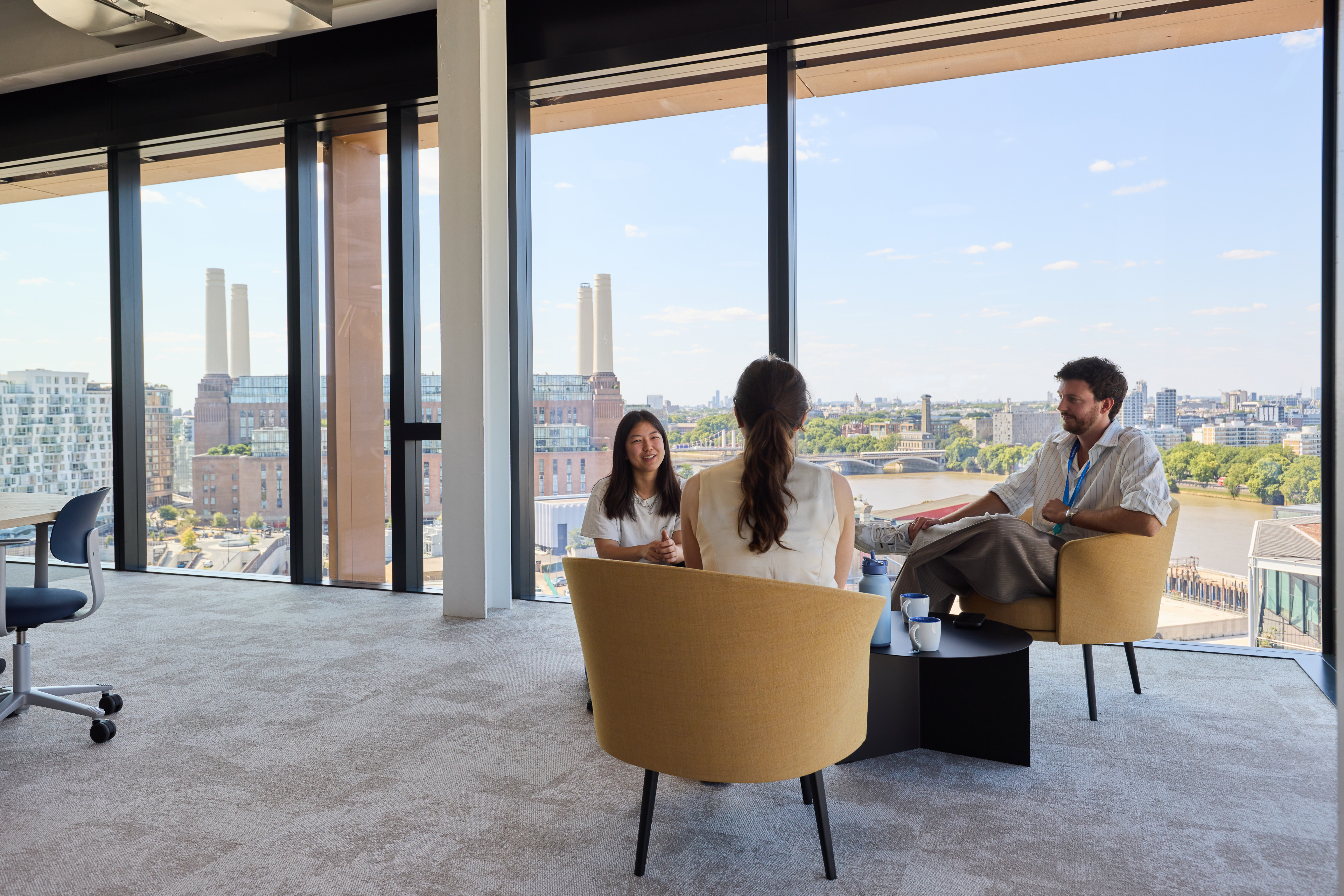 Three people engaged in conversation in a modern office space with large windows overlooking a cityscape featuring the River Thames and notable buildings.