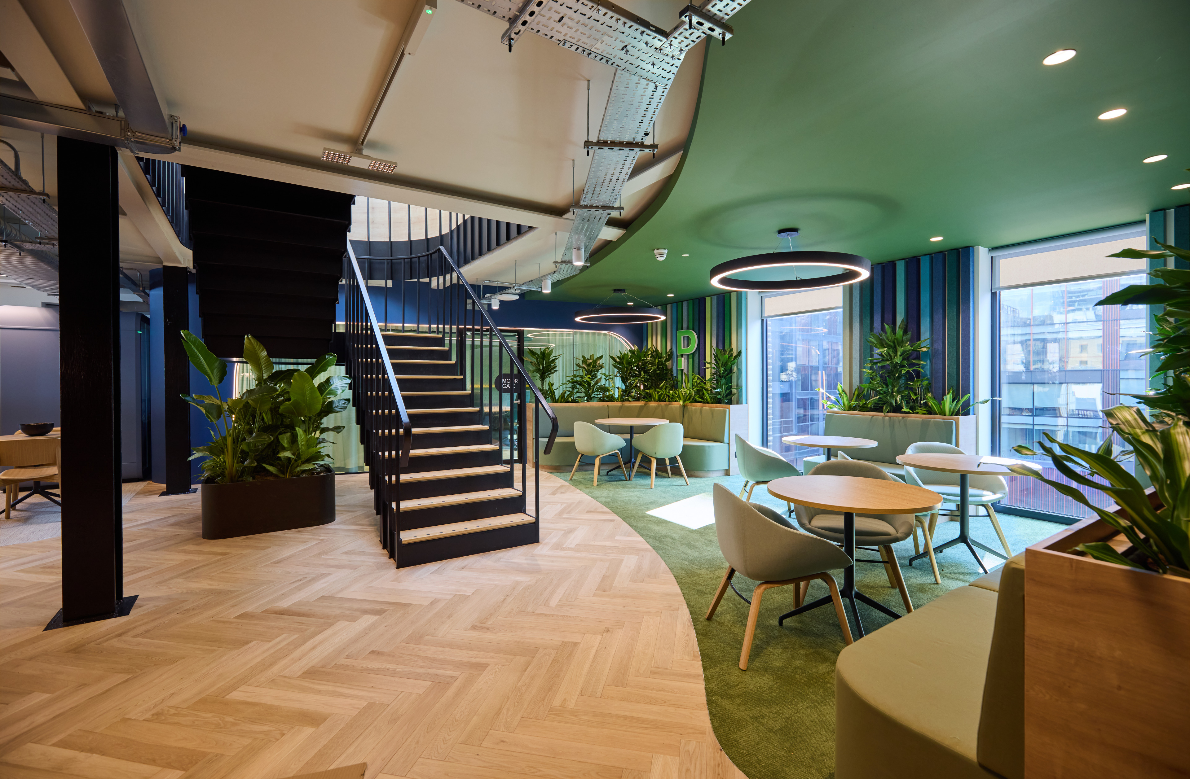 Modern office interior featuring a staircase, seating areas, and greenery, with large windows allowing natural light.