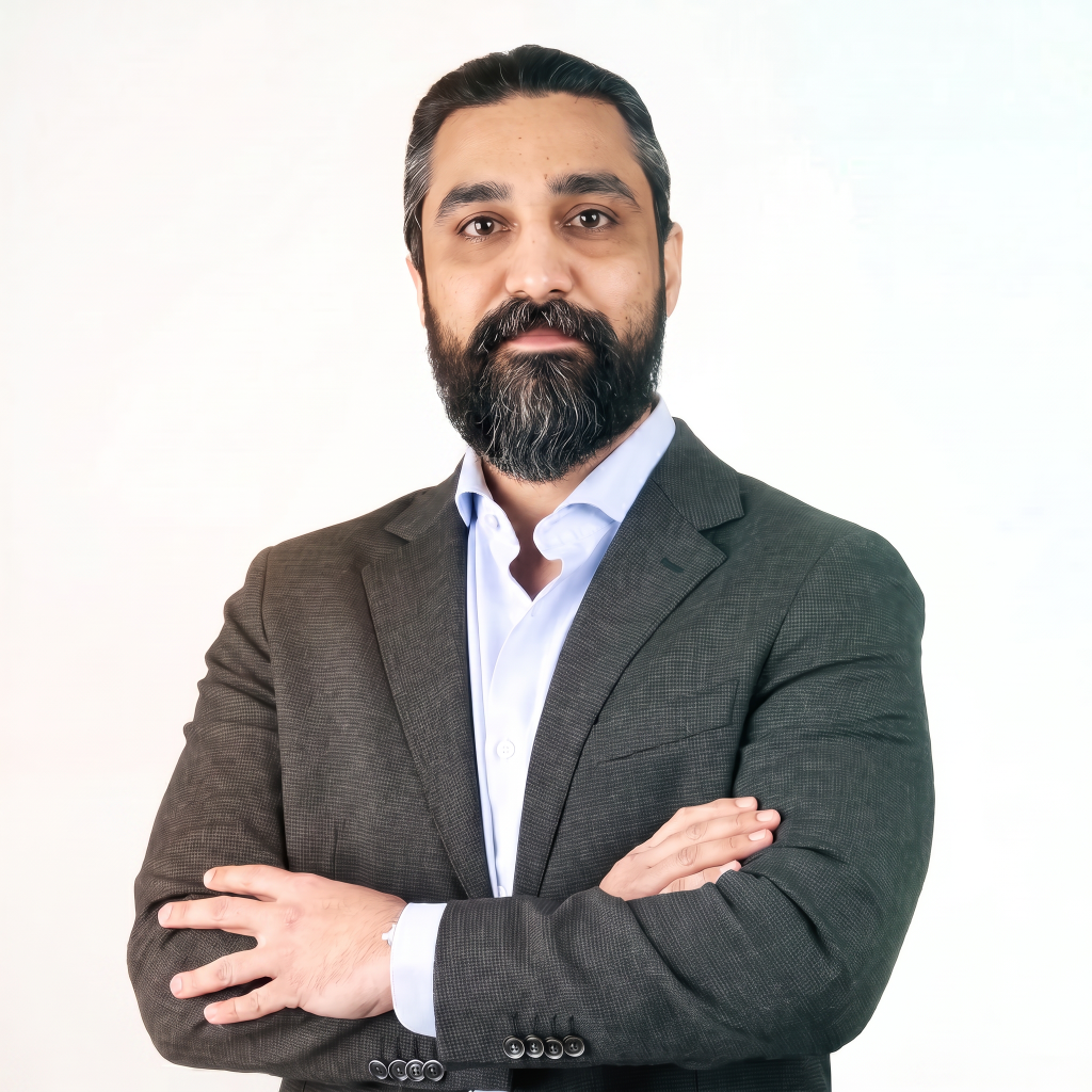 Man with a beard wearing a dark blazer and light shirt, standing with arms crossed against a light background.