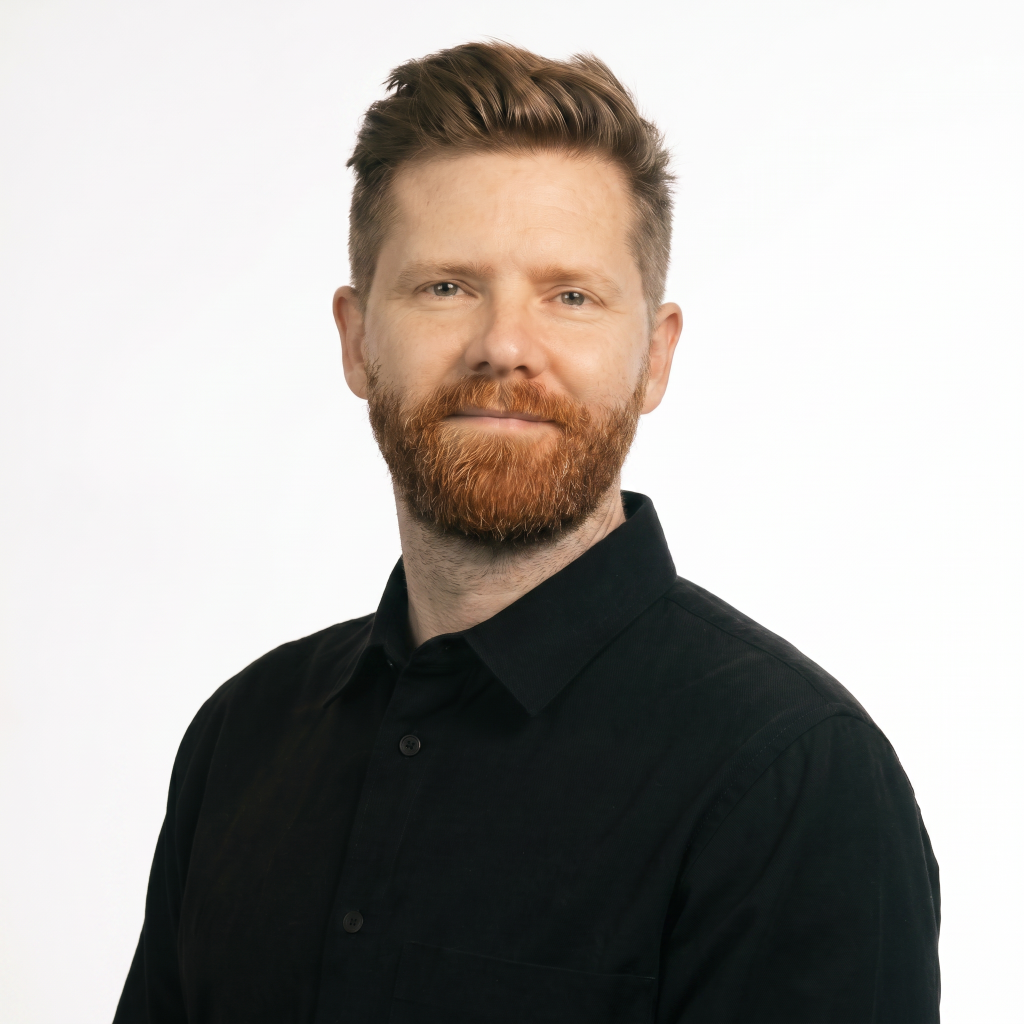Man with a beard and short hair, wearing a black button-up shirt, smiling against a white background.