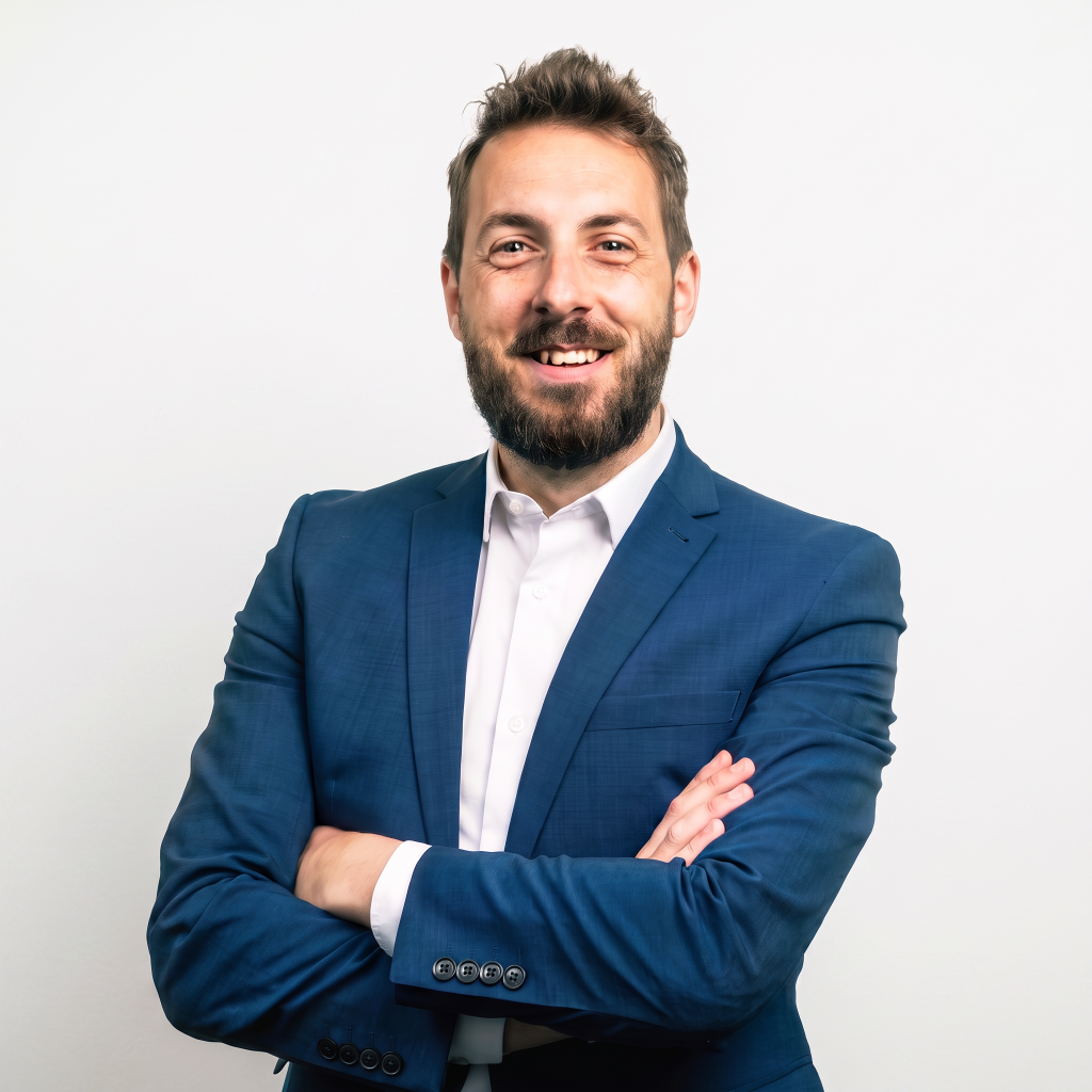 Smiling man with a beard wearing a blue suit and white shirt, standing with arms crossed against a plain white background.