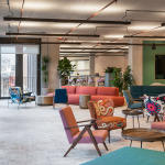 Modern office lounge featuring colorful seating arrangements, plants, and a large TV screen on a green wall.