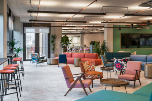 Modern office lounge featuring colorful seating arrangements, plants, and a large TV screen on a green wall.