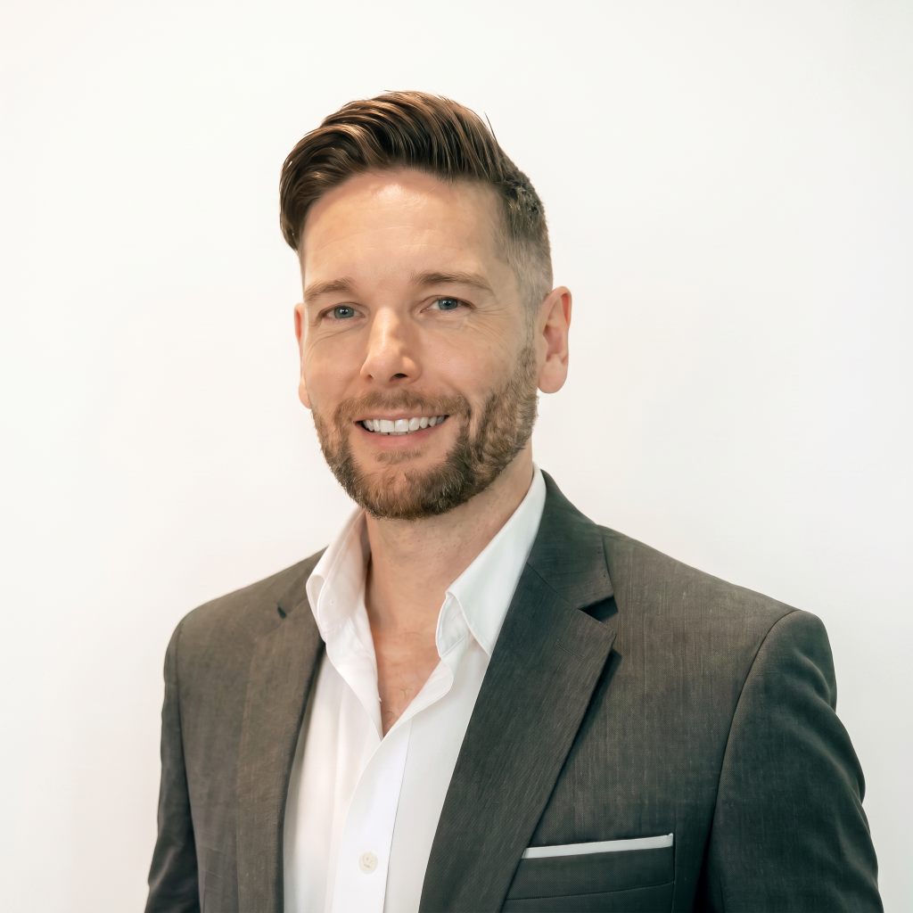 Man with short hair and a beard, wearing a gray suit and white shirt, smiling against a plain white background.