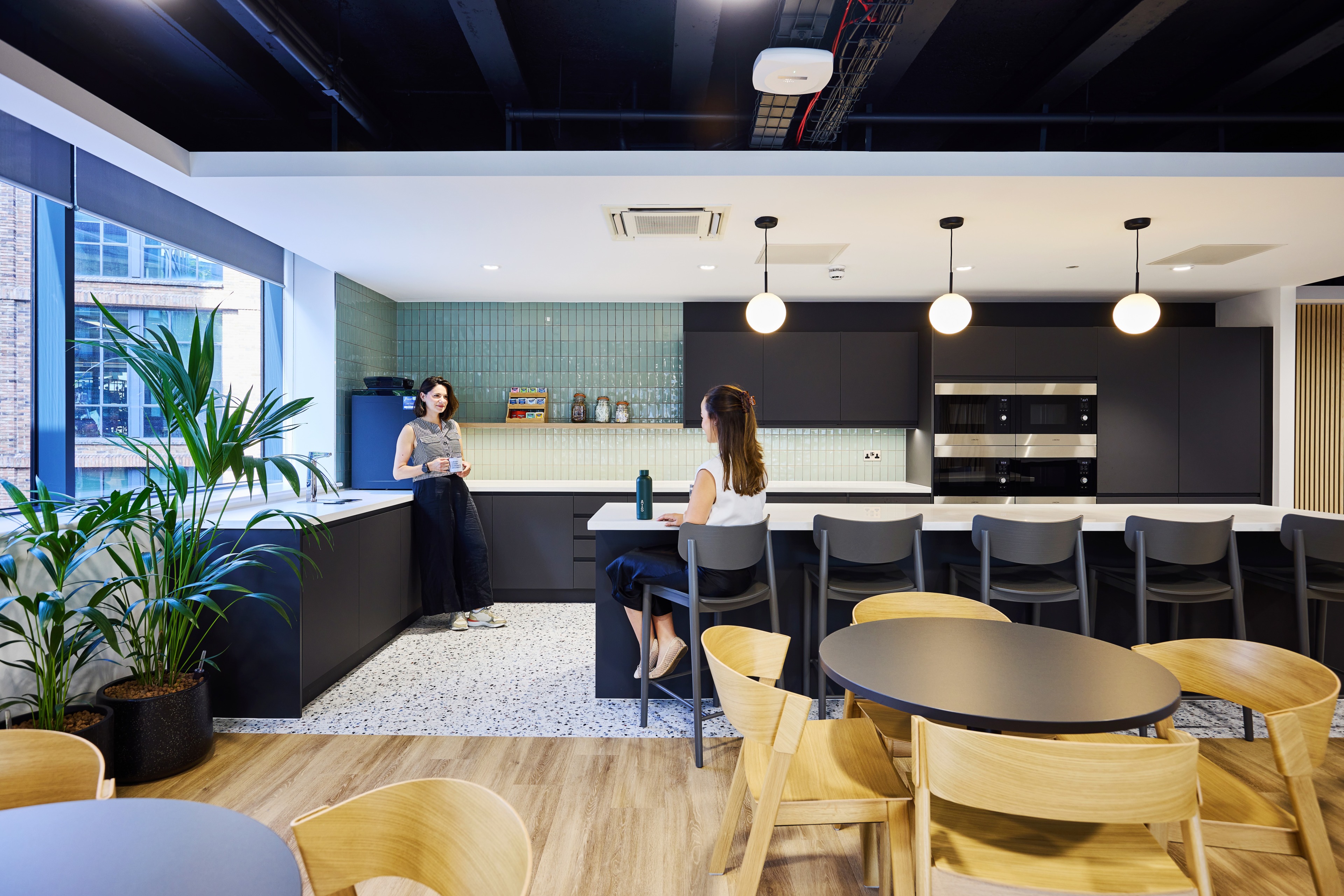 Two women converse in a modern kitchen with green tile accents, a large island, and wooden furniture, while plants are positioned near the windows.